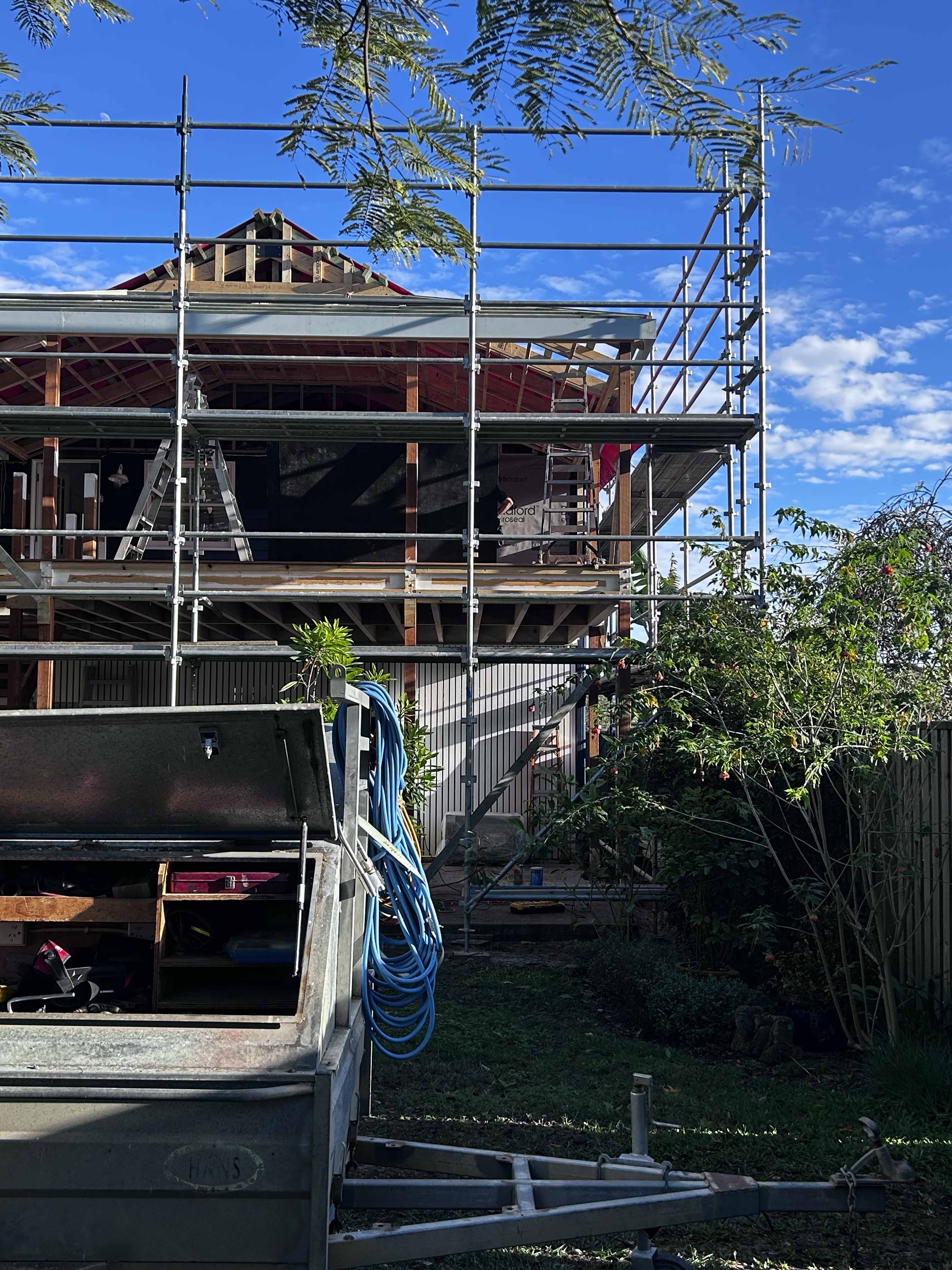 House under construction with scaffolding and garden trailer with tools and coiled blue hose.