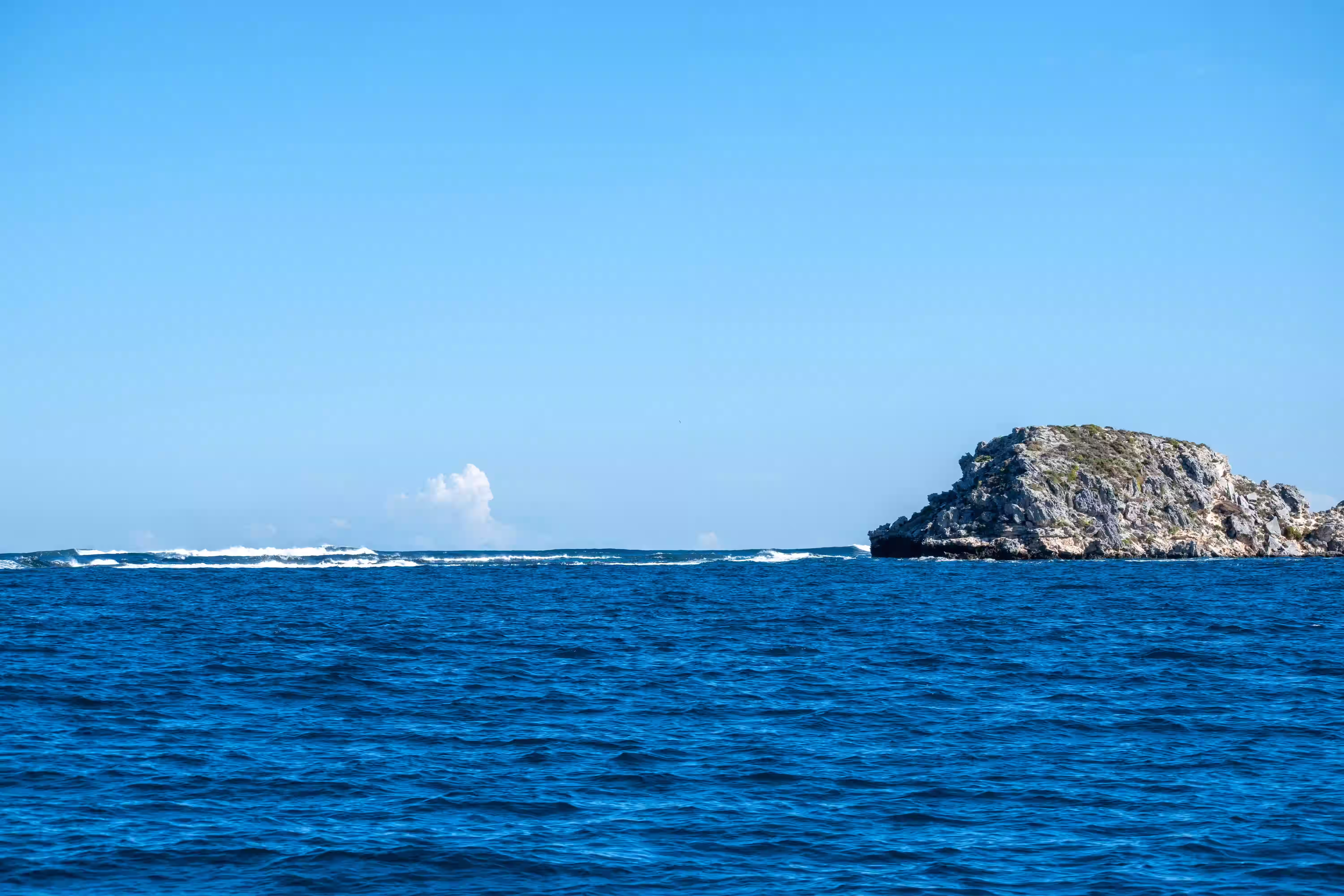 Ocean waves with a rocky island covered in sparse vegetation under a clear blue sky.