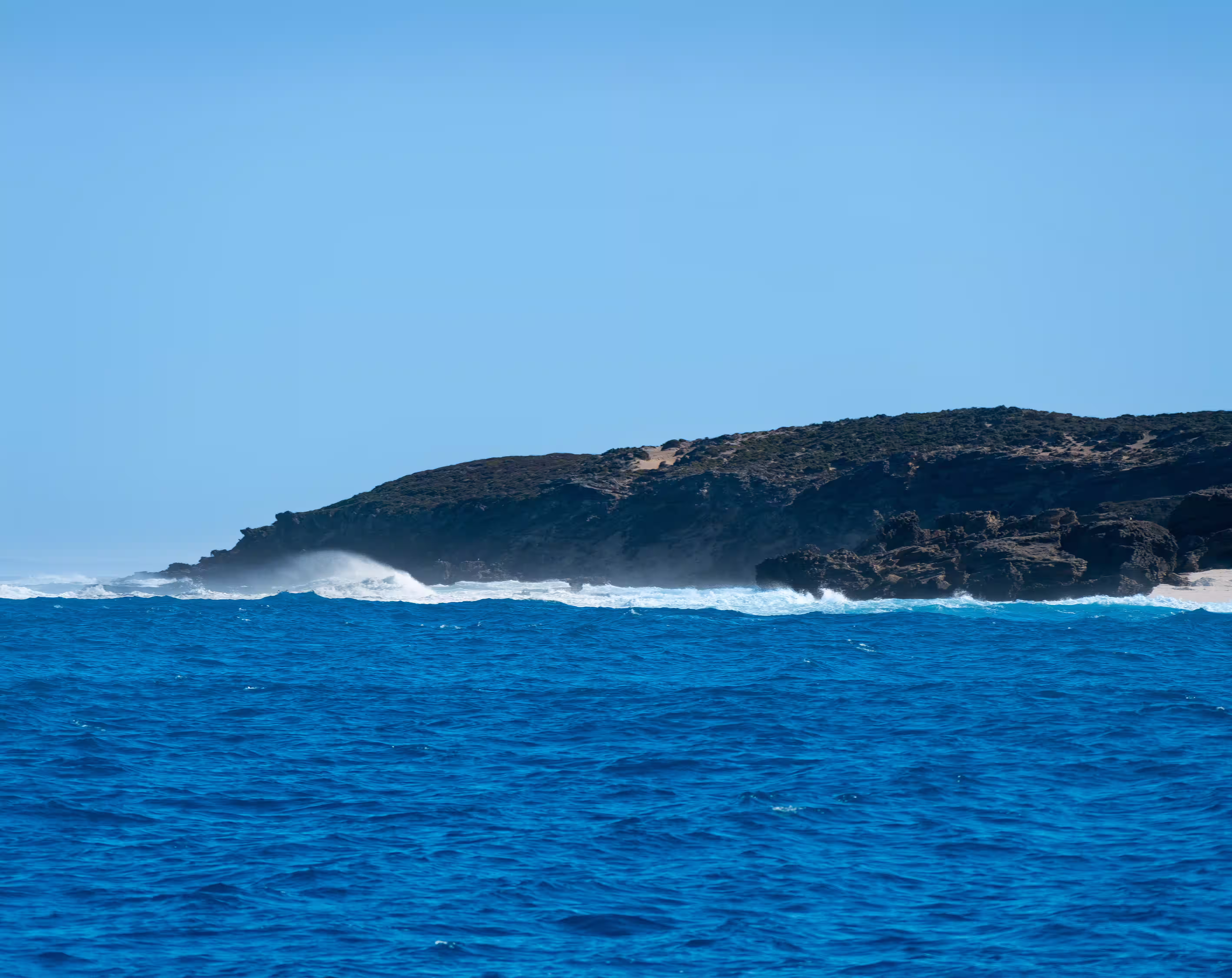 Waves crashing against rocky coastline under a clear blue sky.
