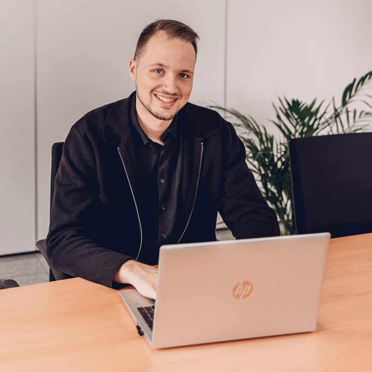 Junger Mann sitzt lächelnd am Tisch und arbeitet an einem silbernen HP-Laptop.
