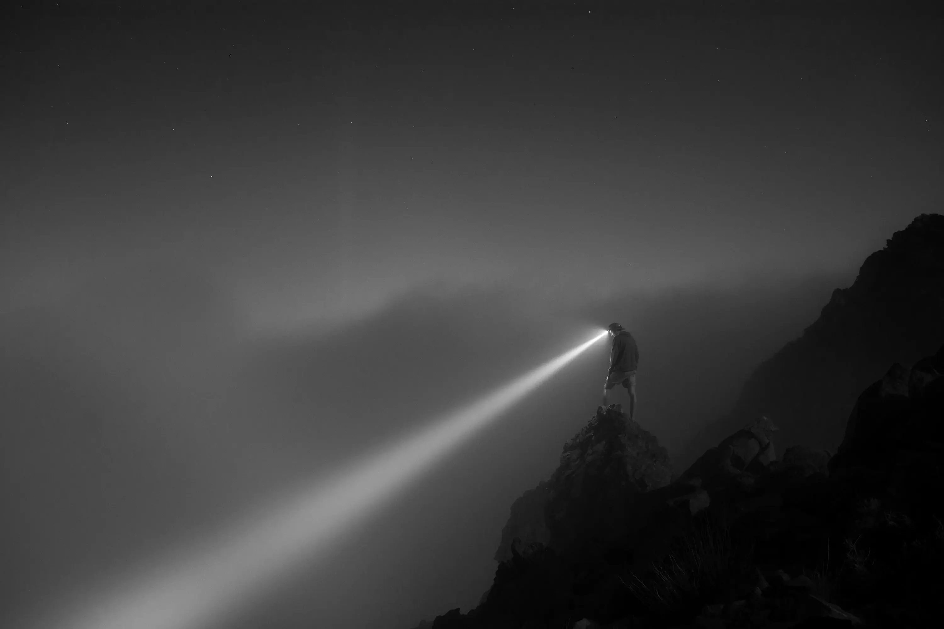 A lone figure on a rocky ridge at night casting a bright headlamp beam into the mist, black and white