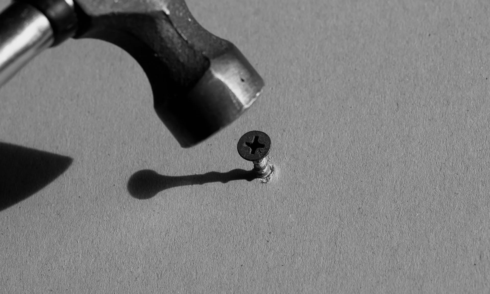 A hammer poised over a screw — the wrong tool for the job — on a grey surface, black and white