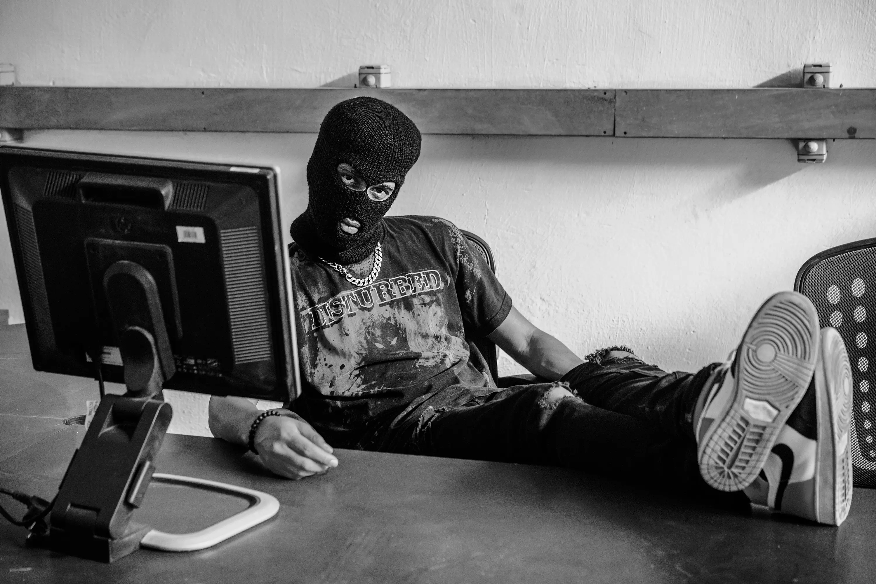 A masked figure in a balaclava slouched at a desk in front of an old computer monitor