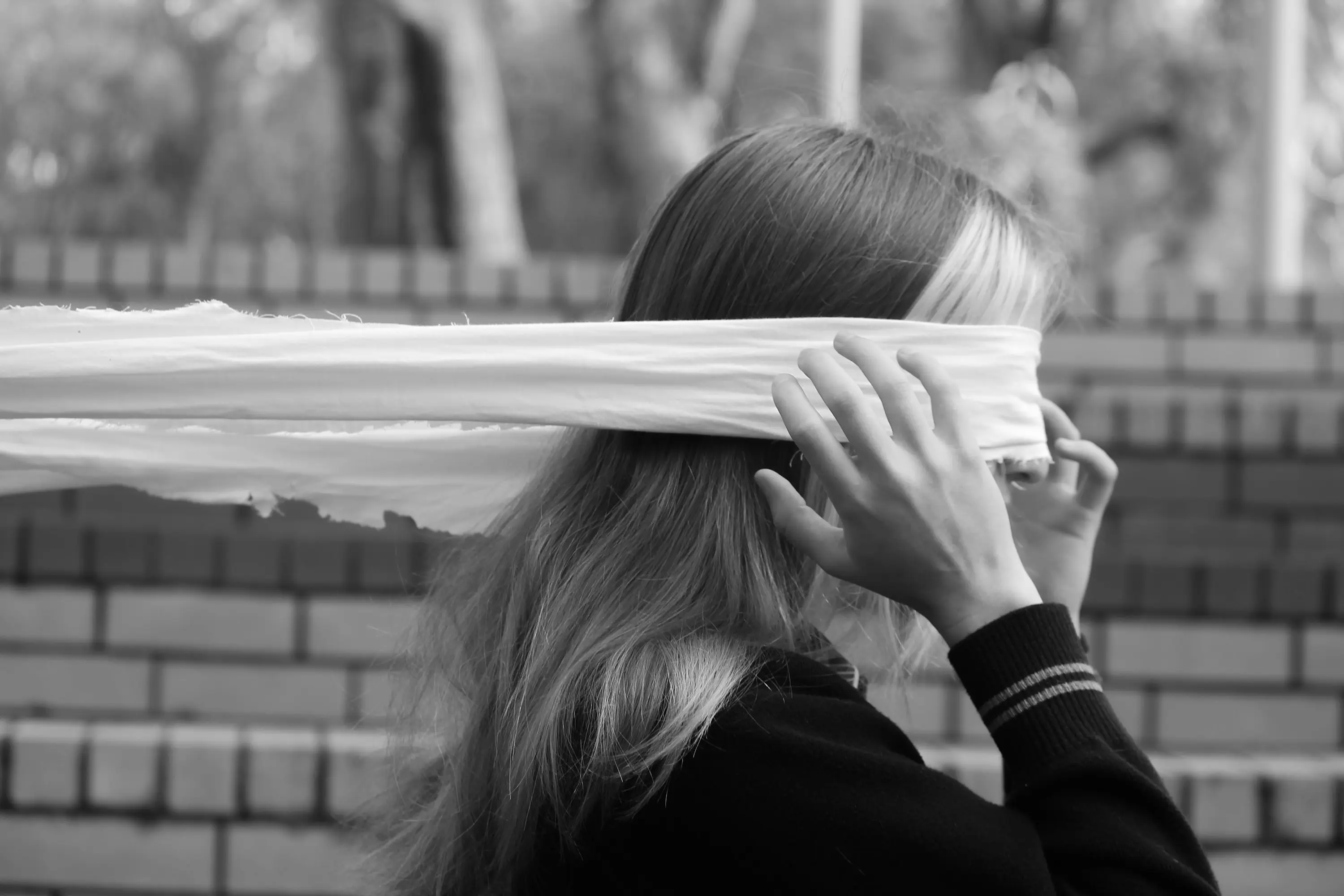 A person pulling a white blindfold across their eyes in front of a brick wall, black and white