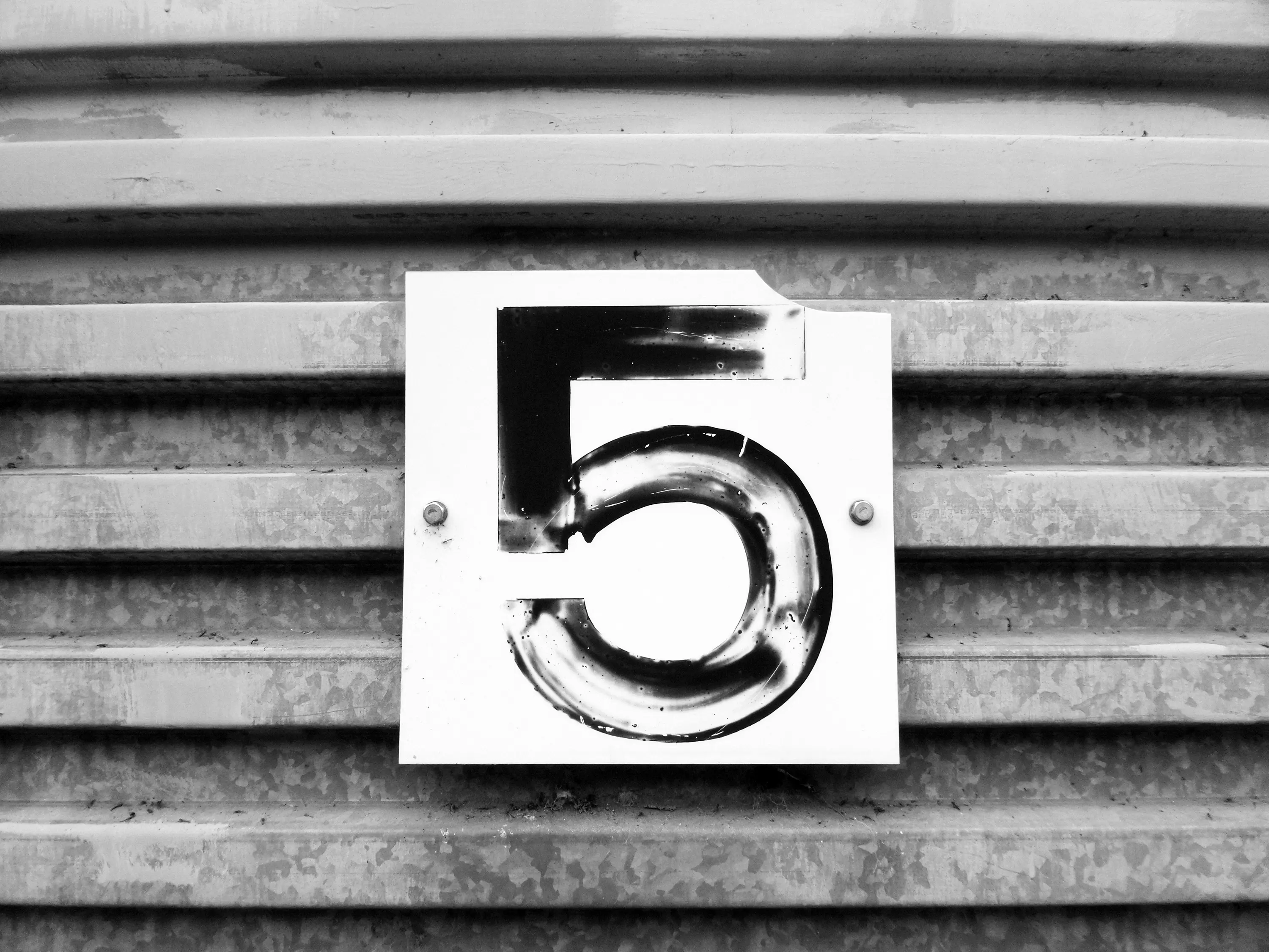 A hand-painted number 5 on a metal plate bolted to a corrugated surface, in black and white
