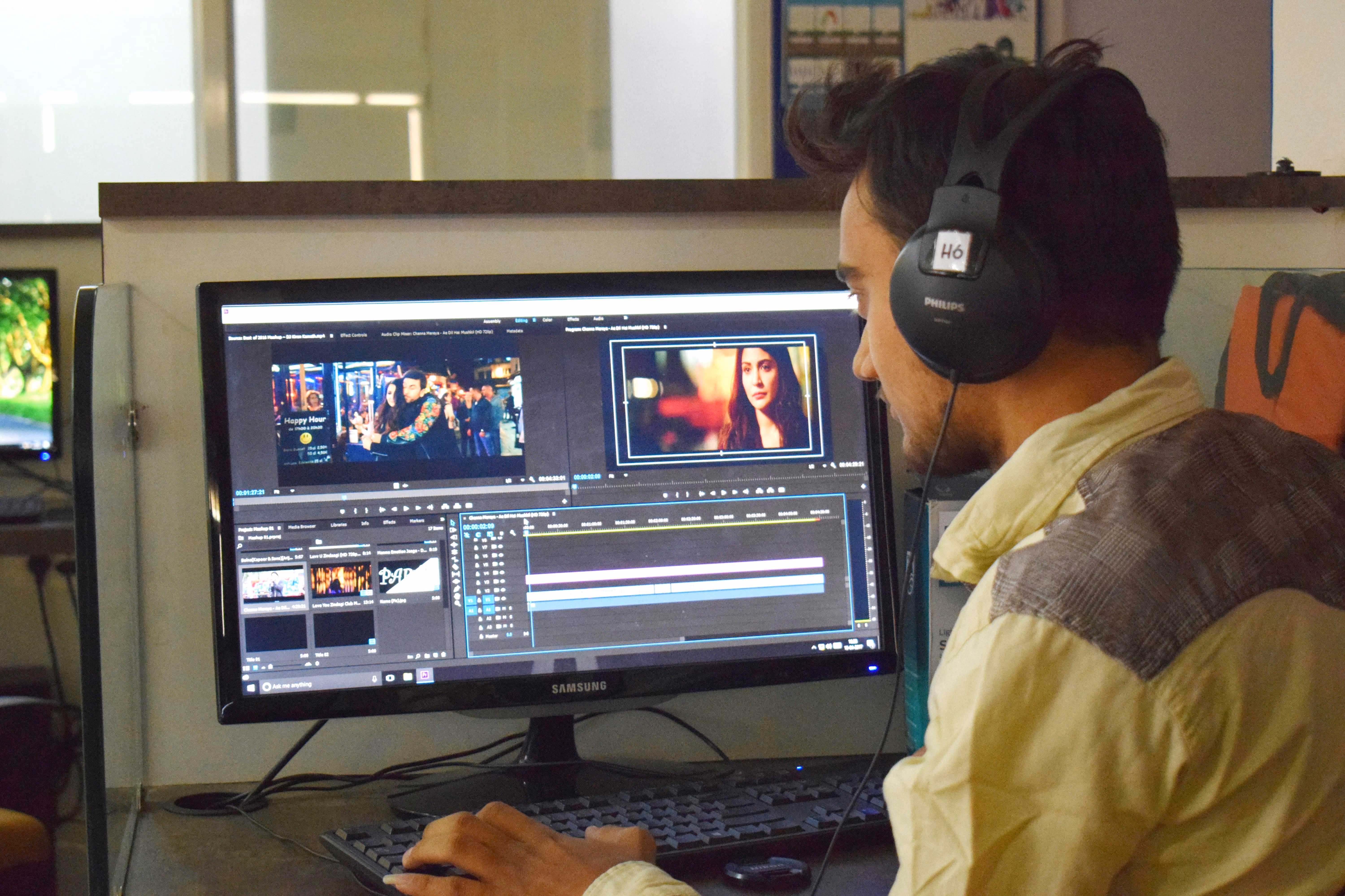 Man wearing headphones working on video editing using software on a Samsung computer monitor in an office setting.