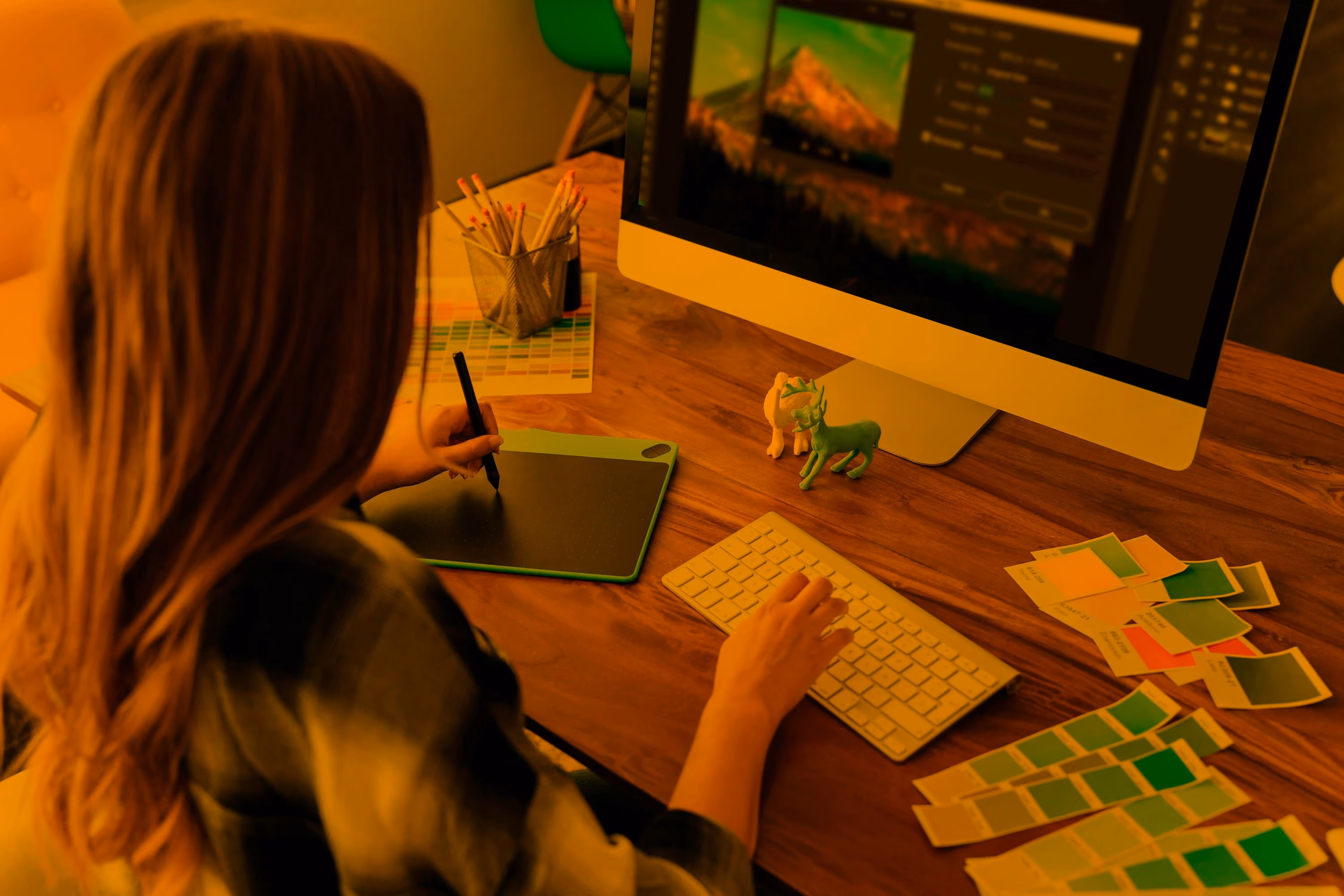 A woman with long hair working at a wooden desk, using a graphics tablet with a stylus in her right hand and a keyboard with her left hand, focused on an iMac screen displaying an image editing software with a mountain photo, surrounded by paint color swatches and pencils.