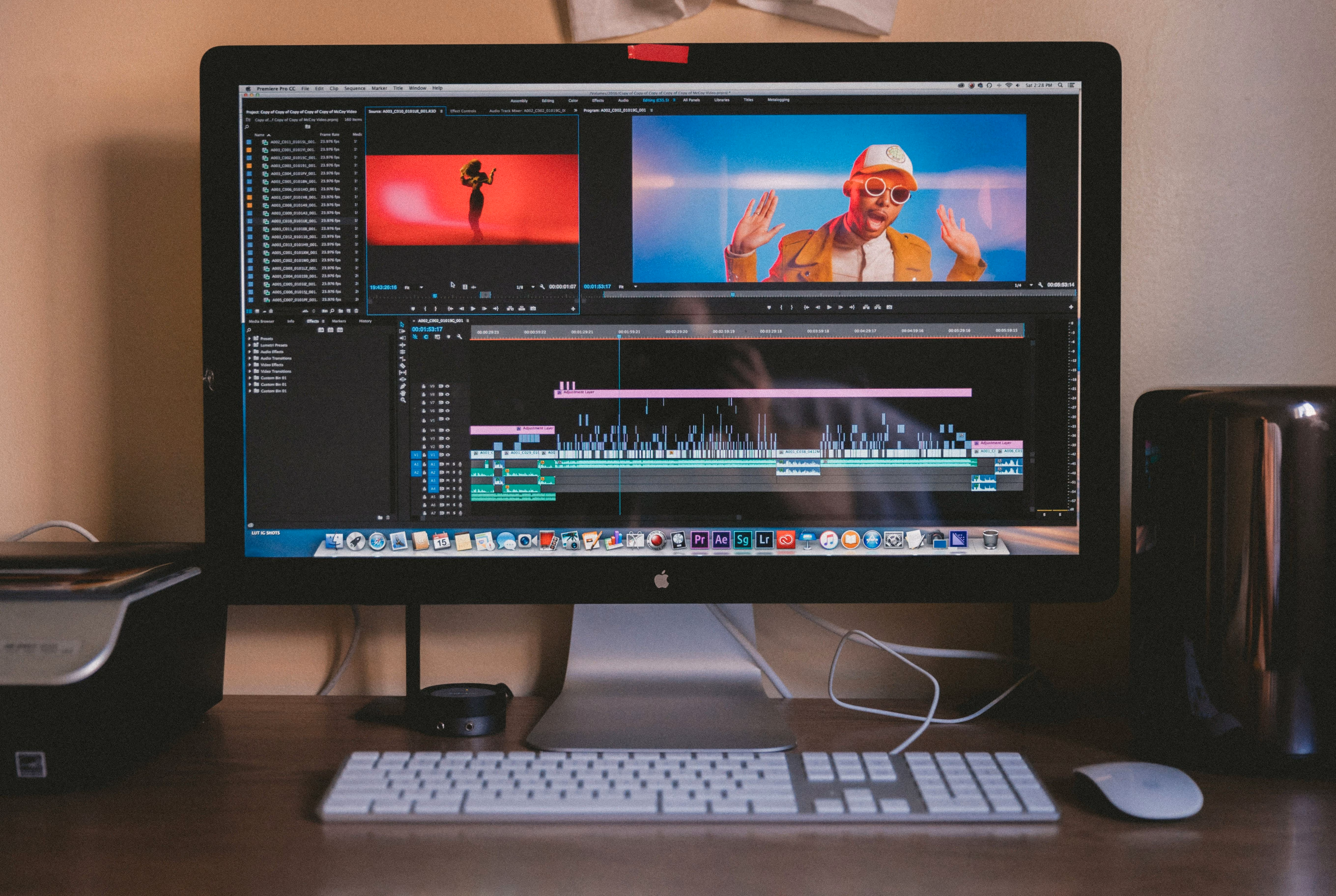 Apple desktop computer on a wooden desk showing video editing software with a timeline, preview windows displaying a person dancing and another person wearing sunglasses and a hat, with keyboard and mouse in front.