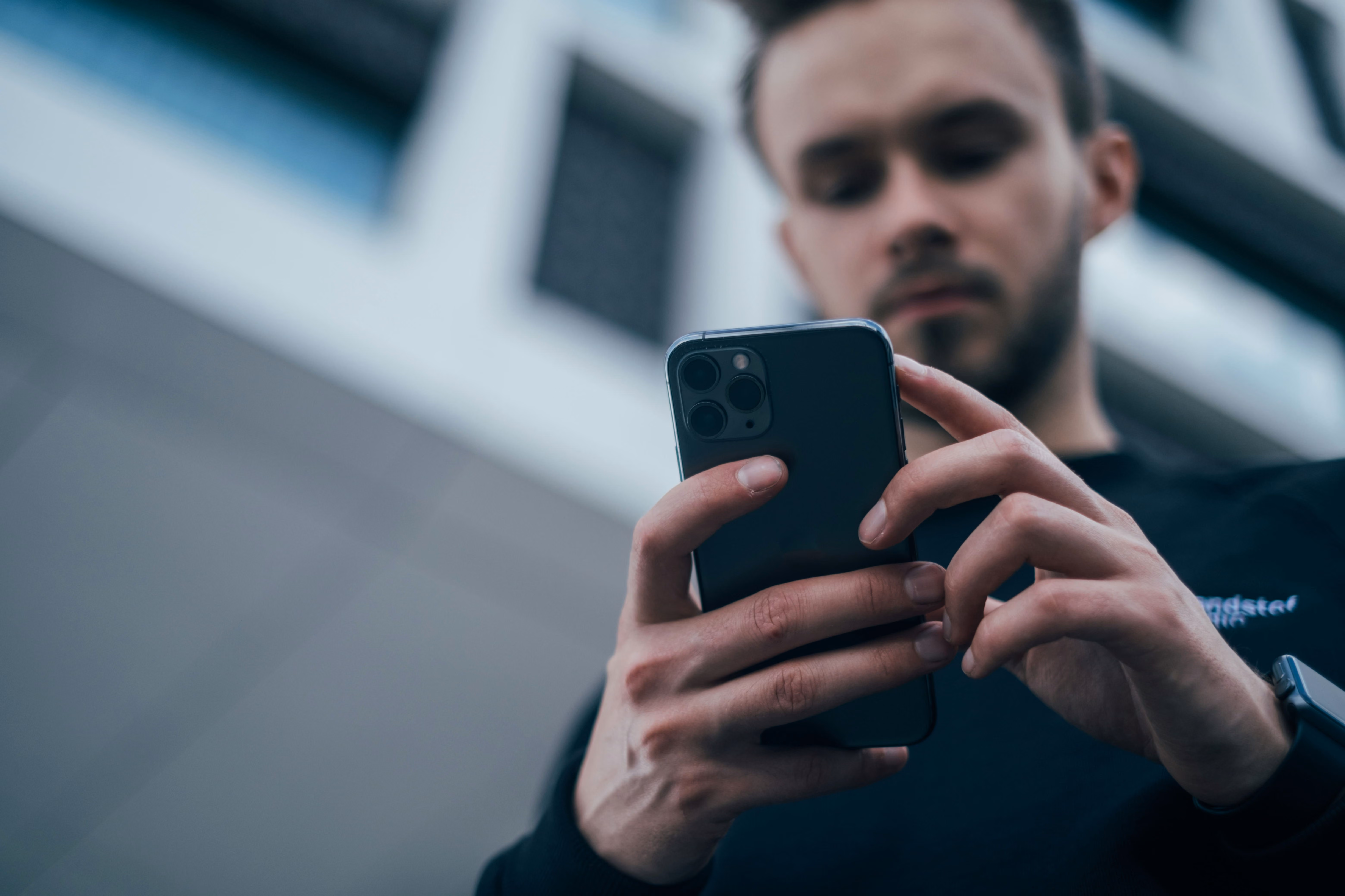 Young man wearing a black shirt using a smartphone with both hands outdoors, with a blurred modern building in the background.