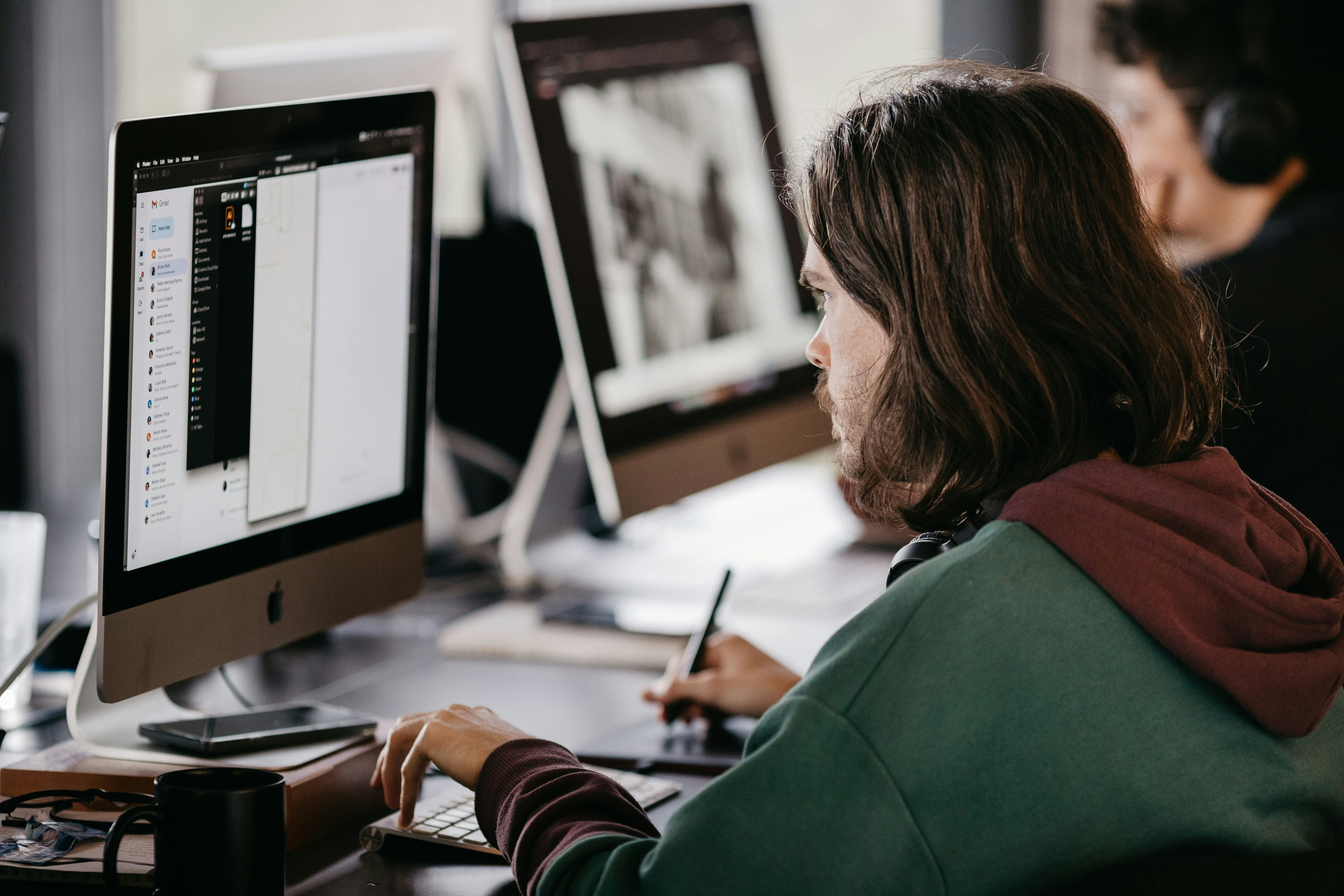 Man with long hair wearing a green and maroon hoodie working on a desktop computer using a stylus and keyboard in an office setting, with another person blurred in the background wearing headphones.