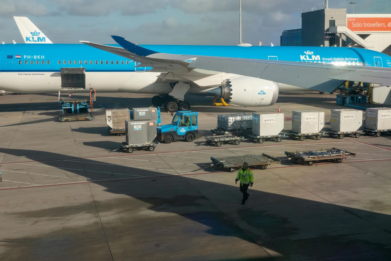 A KLM aircraft at the airport gate undergoing cargo loading and ground handling operations. Containers marked with airline logos are positioned for loading, with the ground crew actively managing logistics.