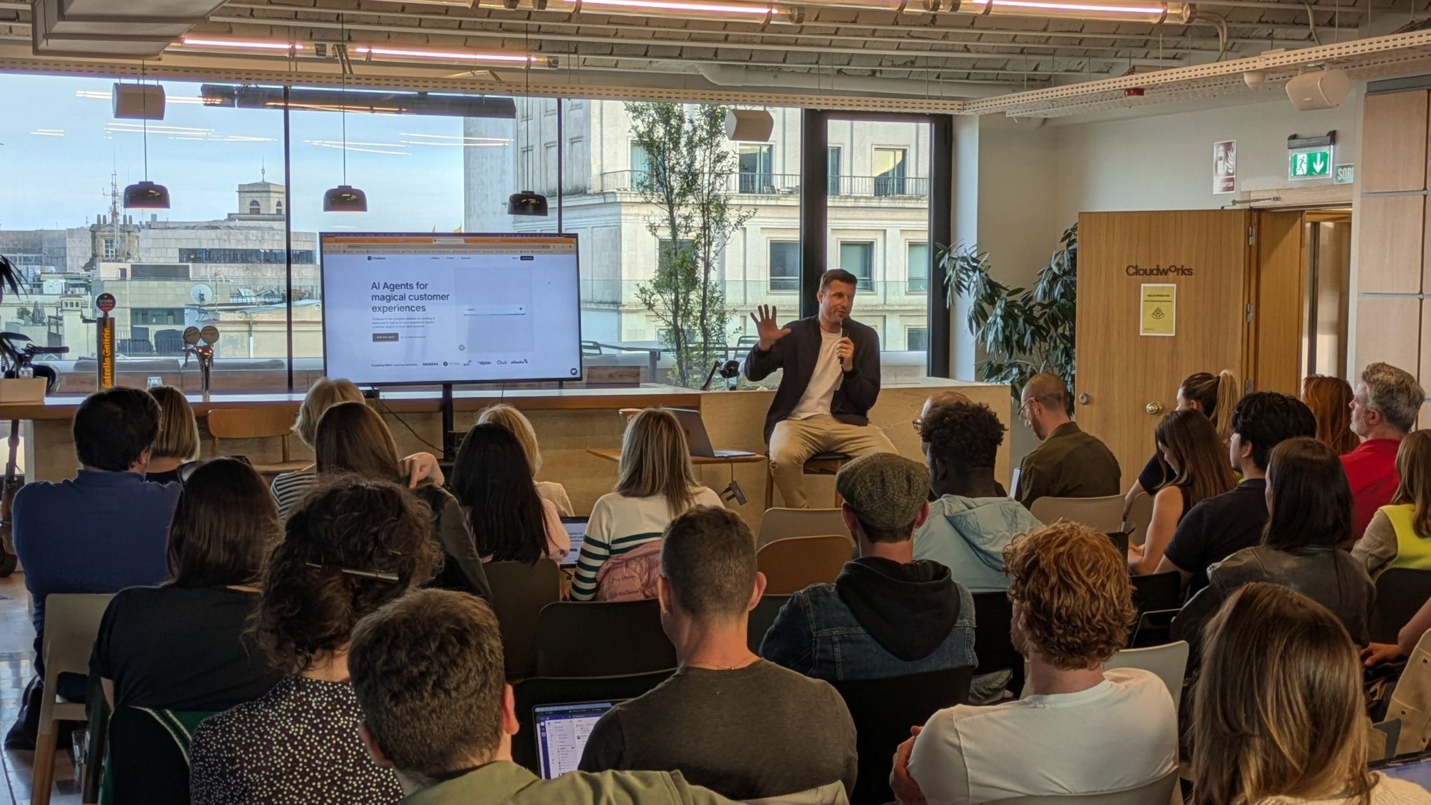 A Hello Operator AI workshop in progress: the speaker, holding a microphone, talks to attendees with laptops open, in front of a screen showing a slide about AI agents.