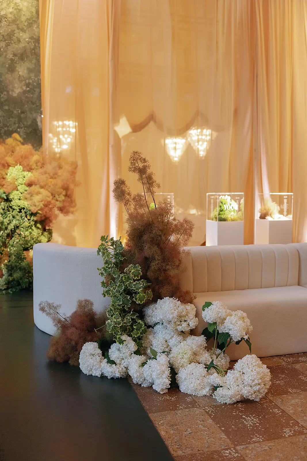 Curved off-white couch beside a cluster of white hydrangeas, green and brown foliage, set on a tiled and dark floor against sheer golden curtains with lit display cases behind.