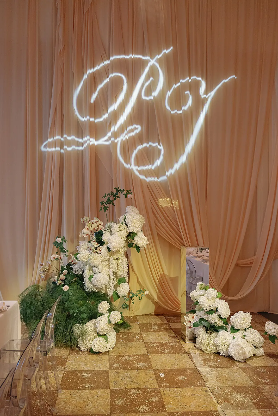 Elegant event setting with a light peach draped backdrop, illuminated white cursive initials projected above, surrounded by white hydrangeas, orchids, greenery, and clear chairs on a tiled floor.