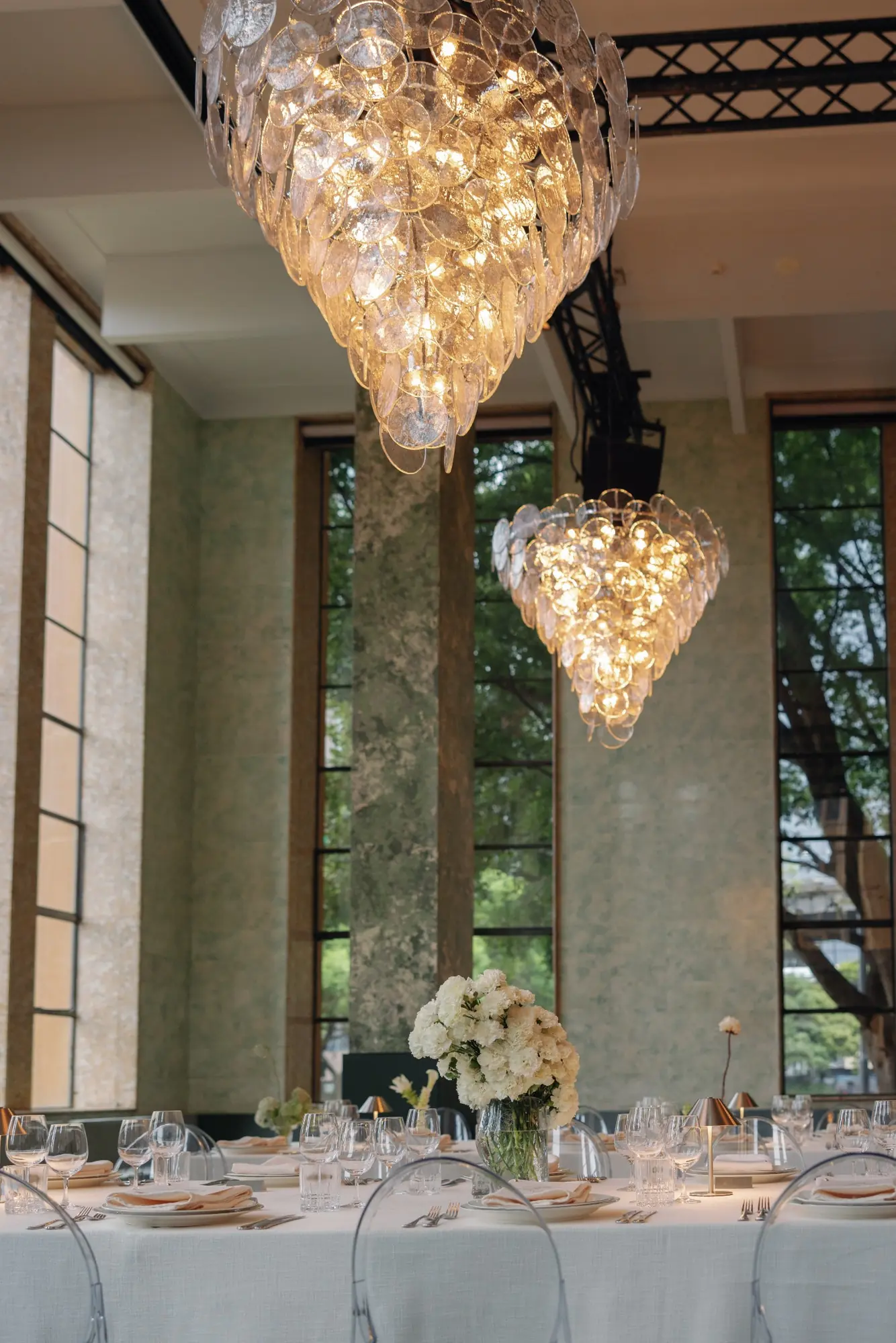 Elegant dining table set with white linens, glassware, silverware, and a vase of white flowers under two large modern chandeliers in a room with tall windows and green walls.