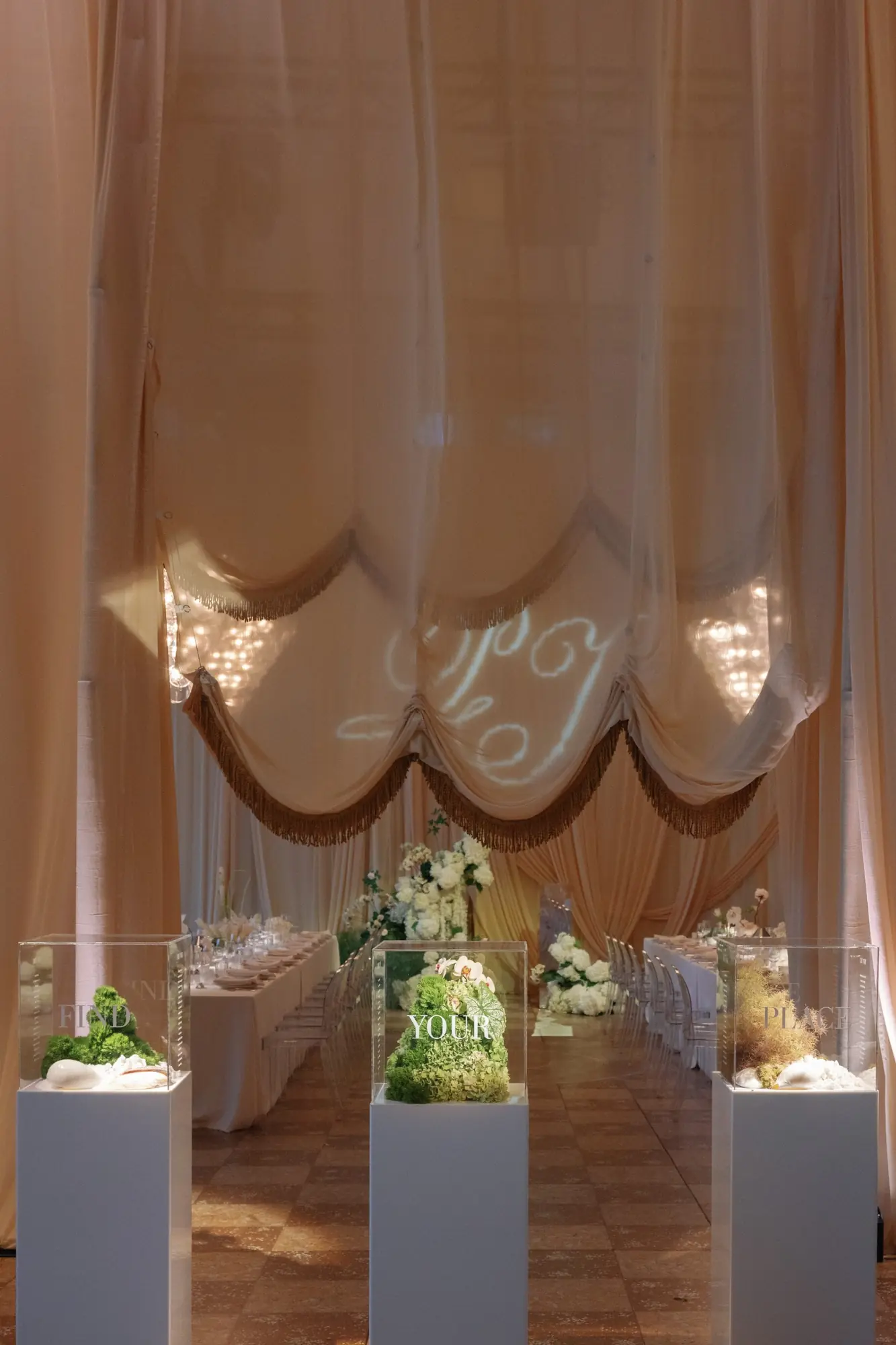Elegant wedding reception setup with long tables, transparent chairs, white floral arrangements, and decorative cubes containing greenery, shells, and the words FIND YOUR PLACE indoors under draped fabric and soft lighting.