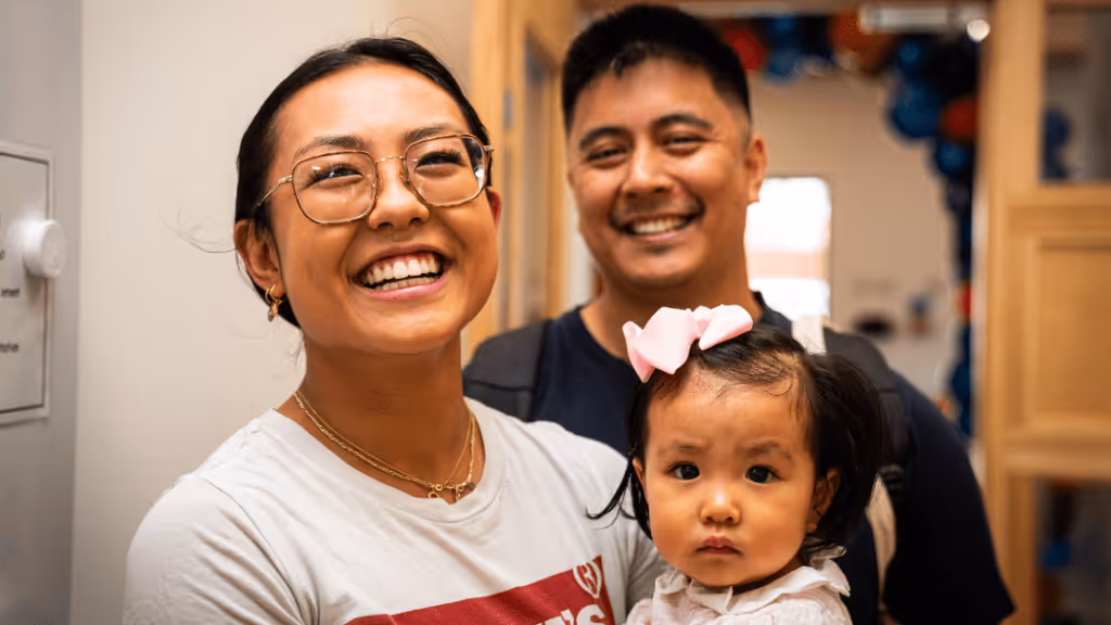 A smiling woman with glasses and a white shirt stands next to a smiling man holding a serious-looking baby girl with a pink bow in her hair. They are indoors, with festive balloons visible in the background.