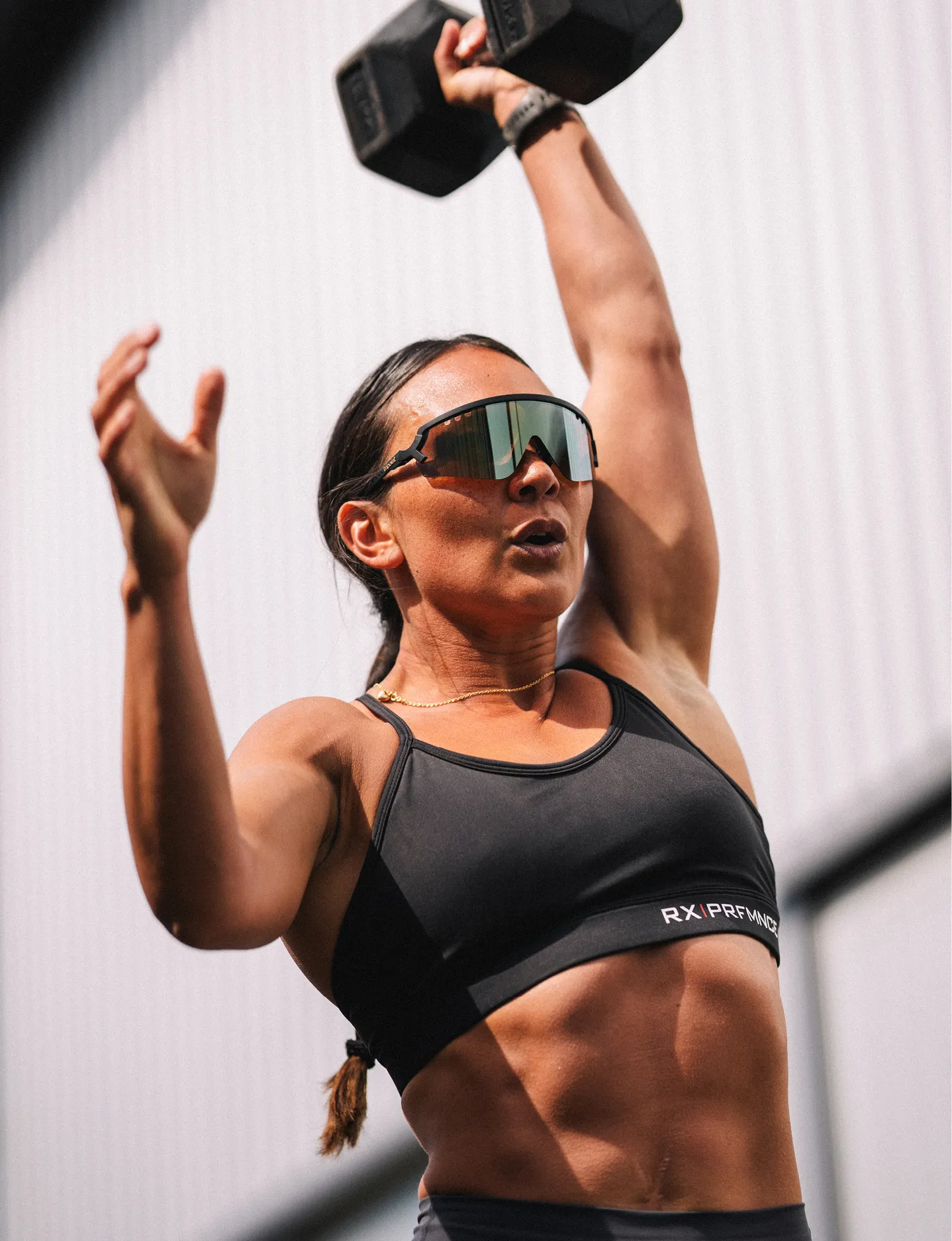 Athletic woman wearing Tripoint sunglasses and a black sports bra lifting a heavy dumbbell overhead during workout.