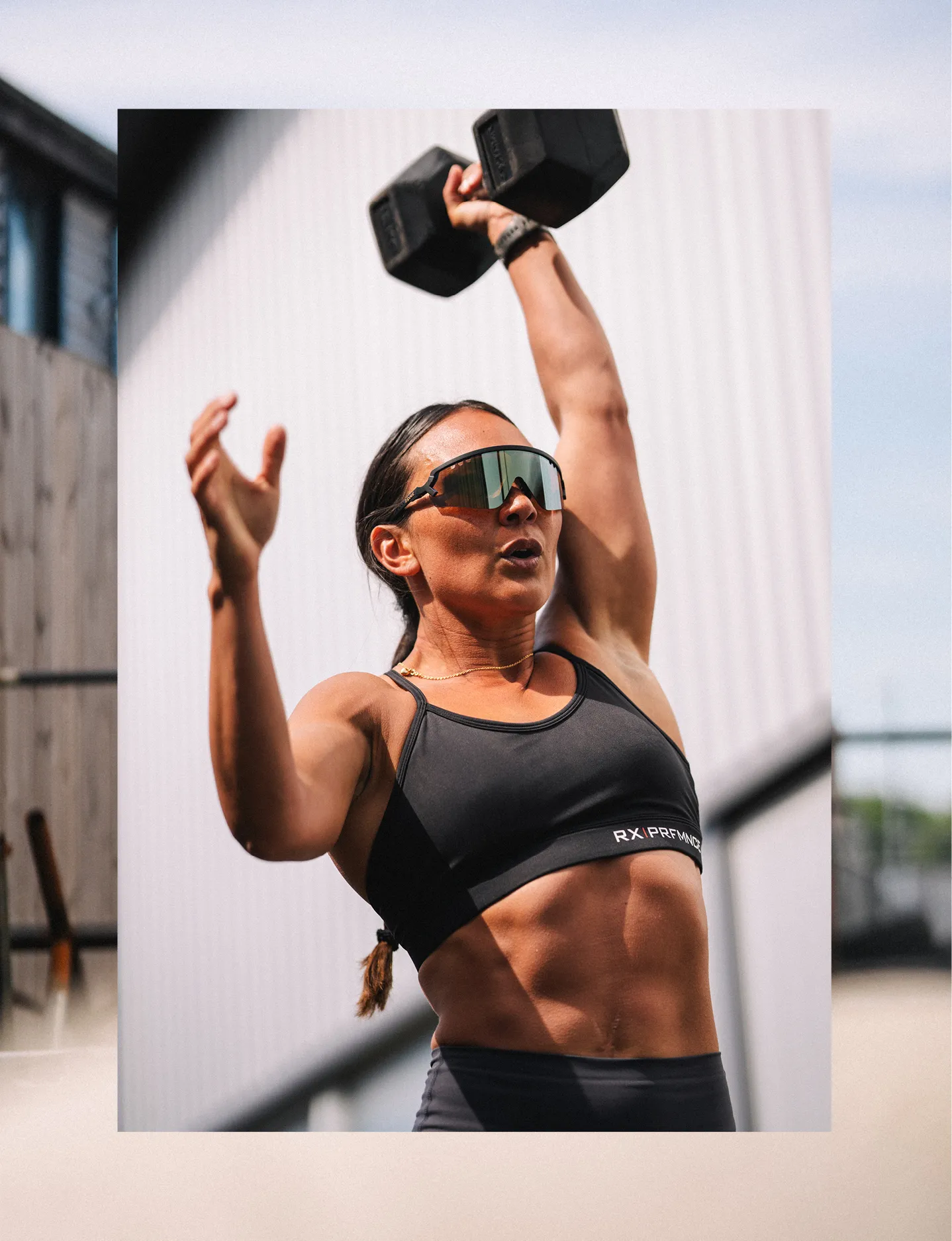Athletic woman wearing black sportswear and Tripoint sunglasses lifting a dumbbell overhead during outdoor workout.
