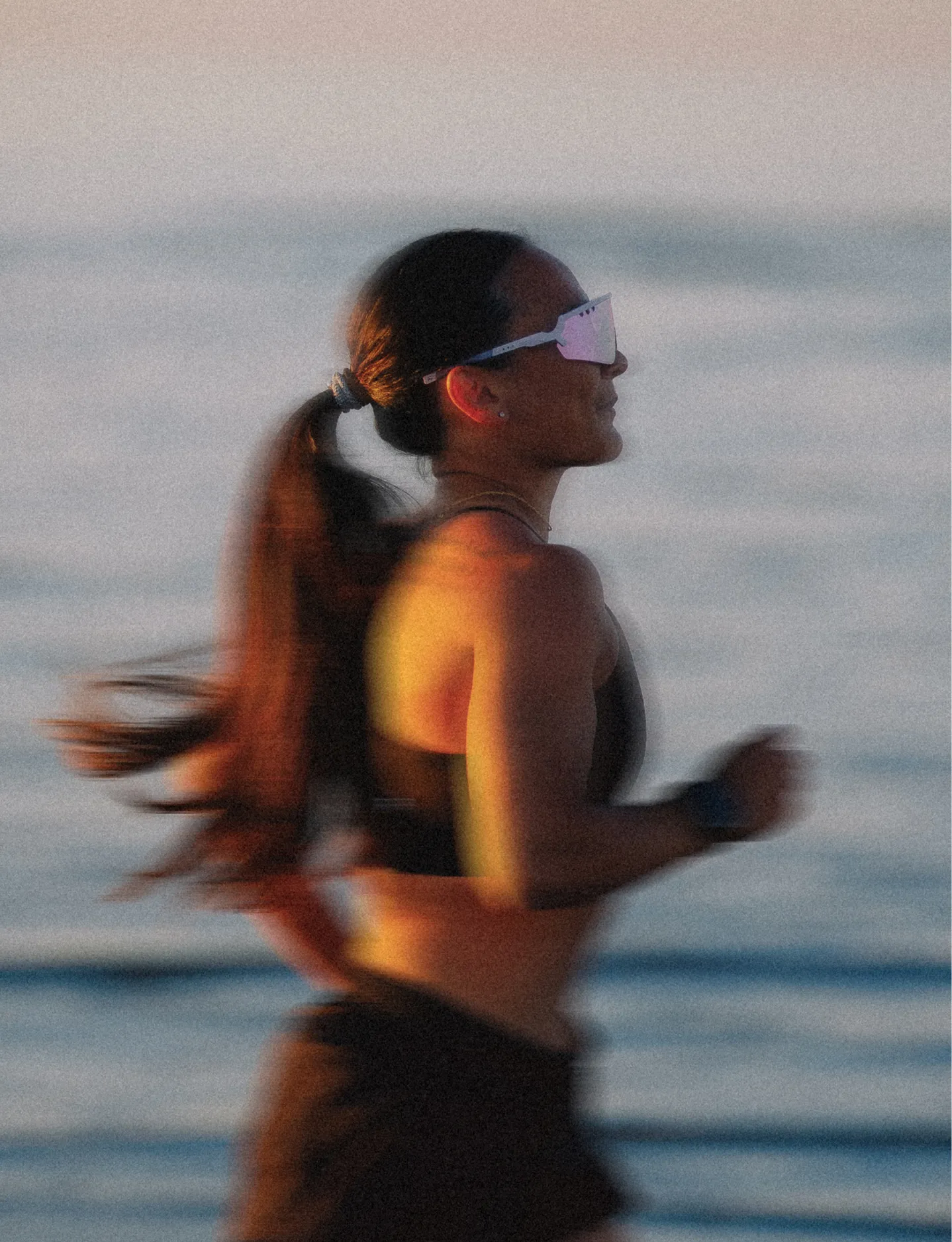 Woman running by the sea in sunset wearing Tripoint sunglasses and a ponytail in soft evening light.