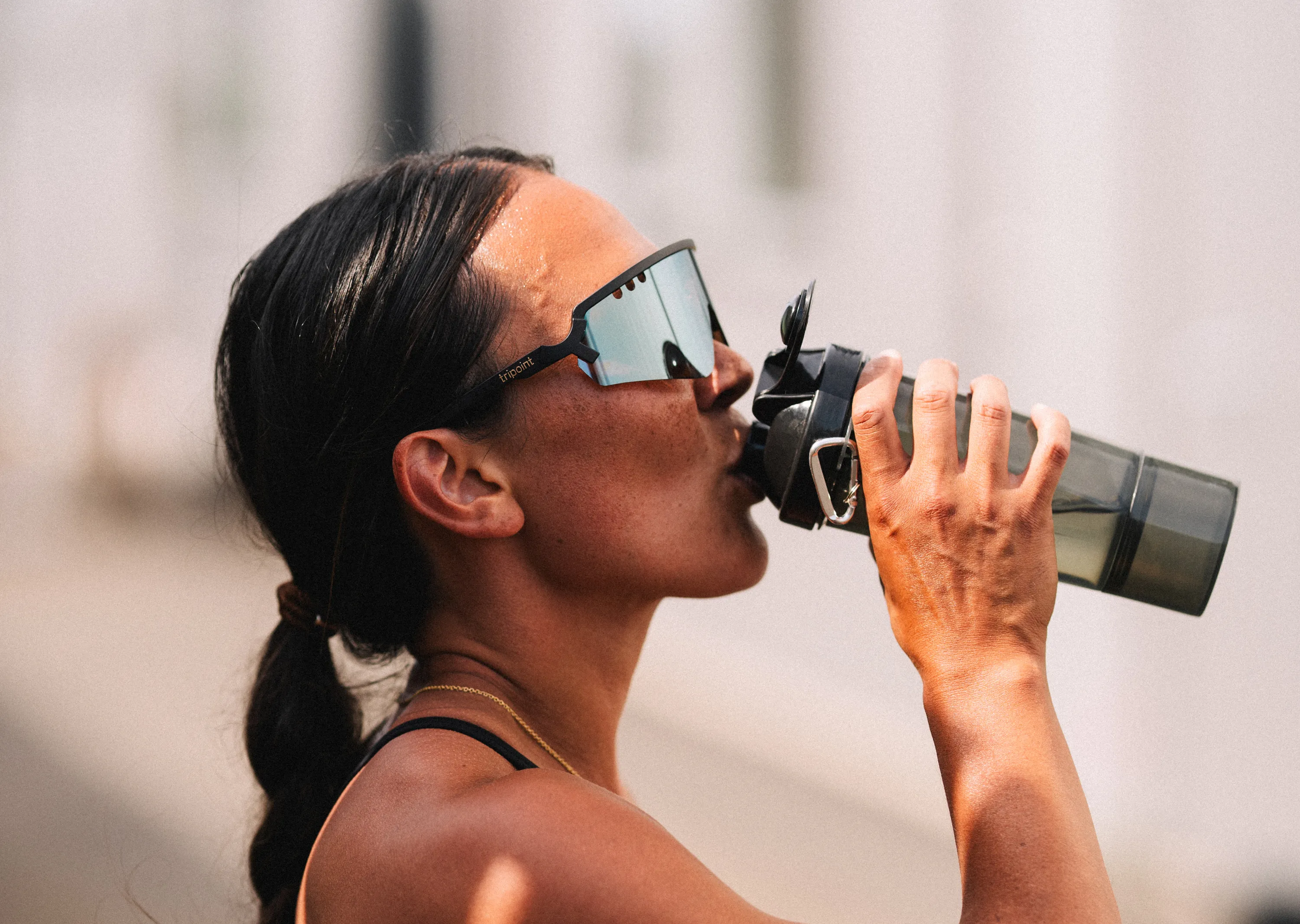 Woman with dark hair tied back wearing mirrored Tripoint sunglasses drinking from a black water bottle.