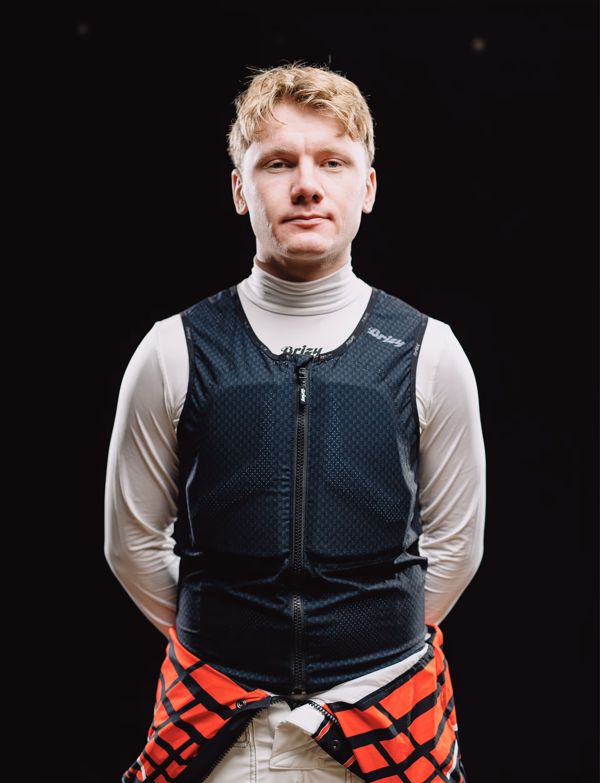 Young man with blonde hair wearing a white turtleneck and a black zip-up vest, standing against a black background.