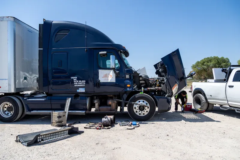 Truck repair on a heavy-duty semi-truck with hood raised as a technician performs engine diagnostics and mechanical repairs beside a mobile service vehicle.