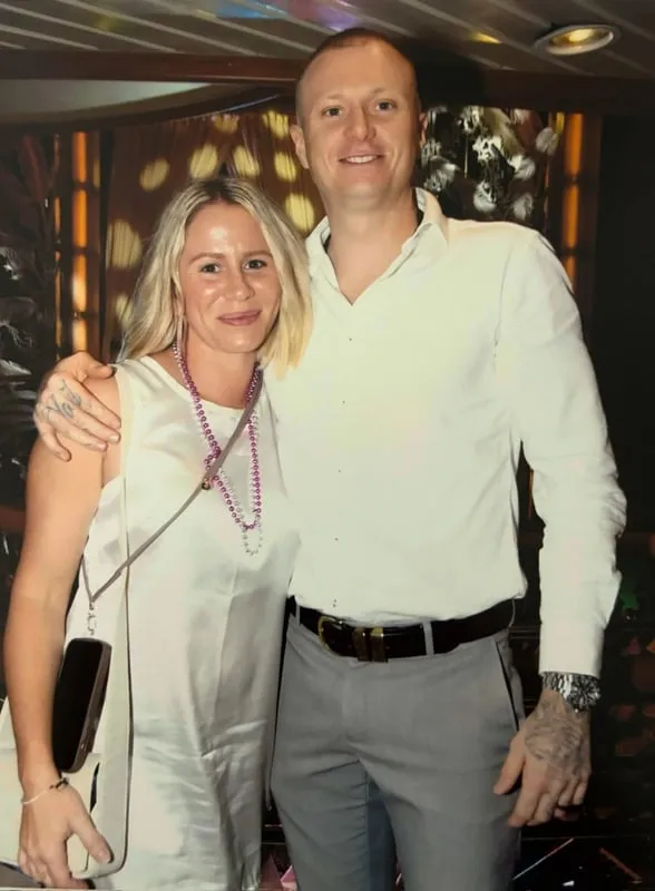 A smiling man in a white shirt with tattoos on his hands and a woman in a white dress with a beaded necklace posing together indoors.