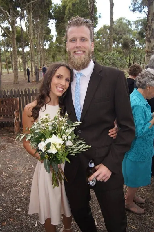 Smiling couple outdoors, woman in a white dress holding a bouquet and man in a suit holding a beer bottle.