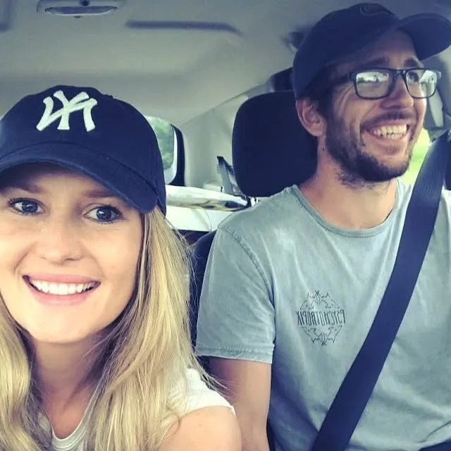 Smiling woman with long blonde hair wearing a black cap with white logo, and a man with glasses and a beard wearing a blue cap and seatbelt inside a car.