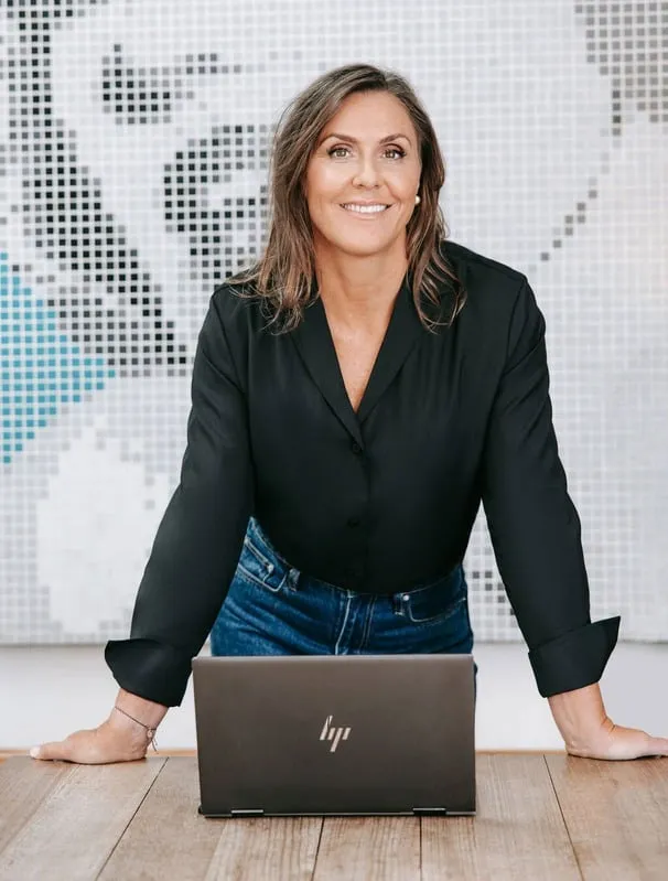 Smiling woman leaning on a wooden table with a laptop in front of her, standing against a pixelated mosaic background.