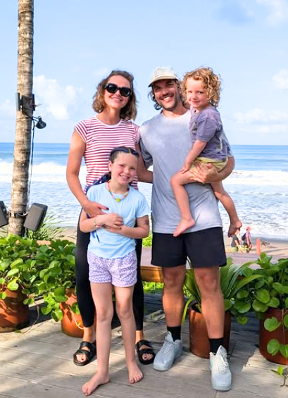 Smiling family of four with two children posing outdoors by the ocean with plants and palm tree in the background.