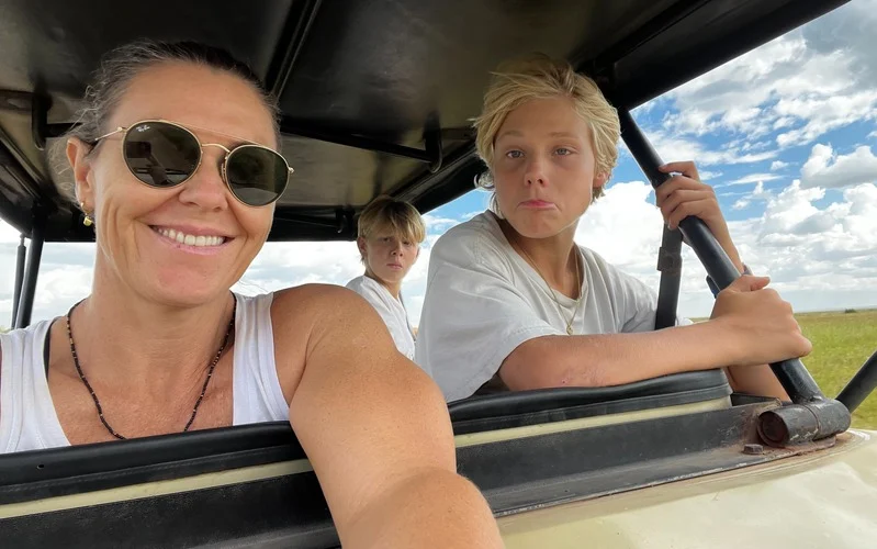 Three people sitting in an open vehicle outdoors under a partly cloudy sky, with a woman wearing sunglasses smiling in the foreground.