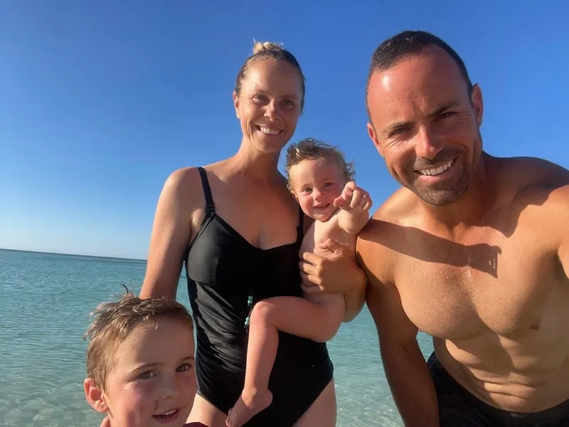 Smiling family of four at the beach with clear blue sky and calm sea in the background.