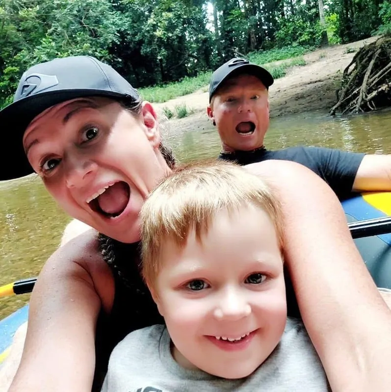 Three people on a blue inflatable raft in a river, smiling and making surprised faces with trees and a dirt riverbank in the background.