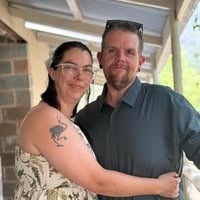 Smiling family of five sitting on a bed with a woman holding a baby, a girl in white, and a man holding a young boy.