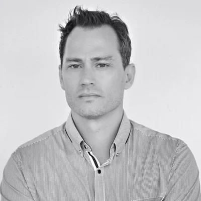 Black and white portrait of a man with short dark hair wearing a striped button-up shirt, looking directly at the camera with a neutral expression.