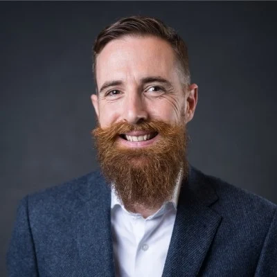 Smiling man with a thick red beard and mustache wearing a gray blazer and white shirt against a dark background.