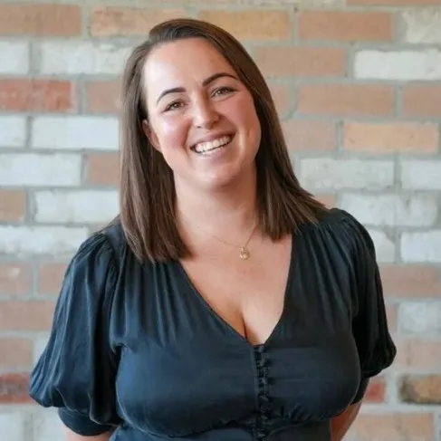 Smiling woman with shoulder-length brown hair wearing a black top standing against a brick wall.