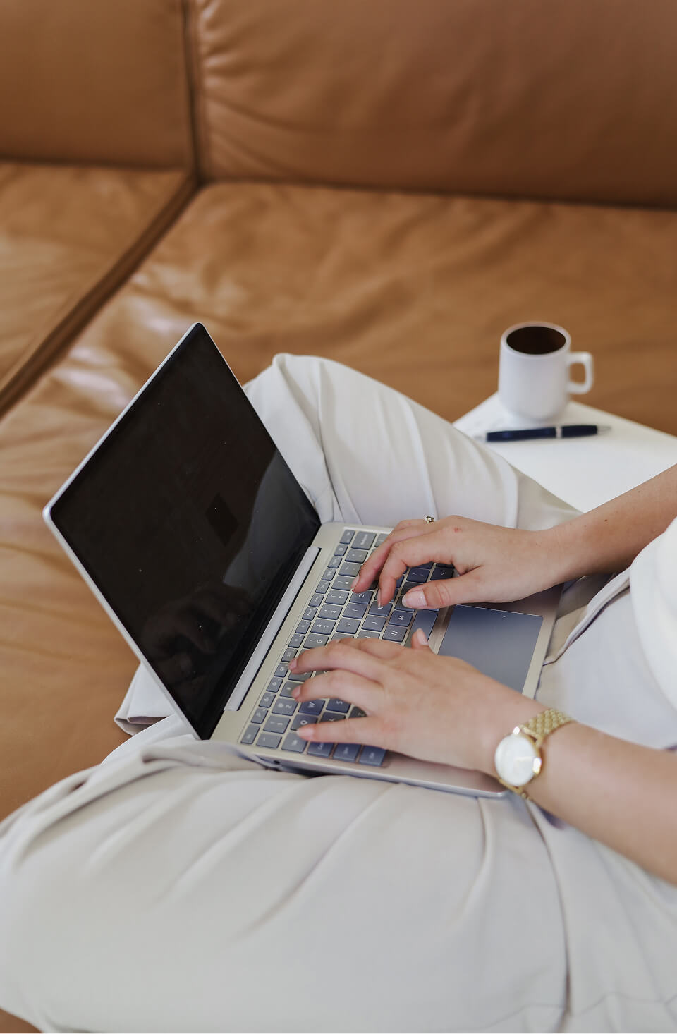 Ashley from Noma Finance typing on a laptop while sitting on a brown leather couch with a white cup and pen on a side table.