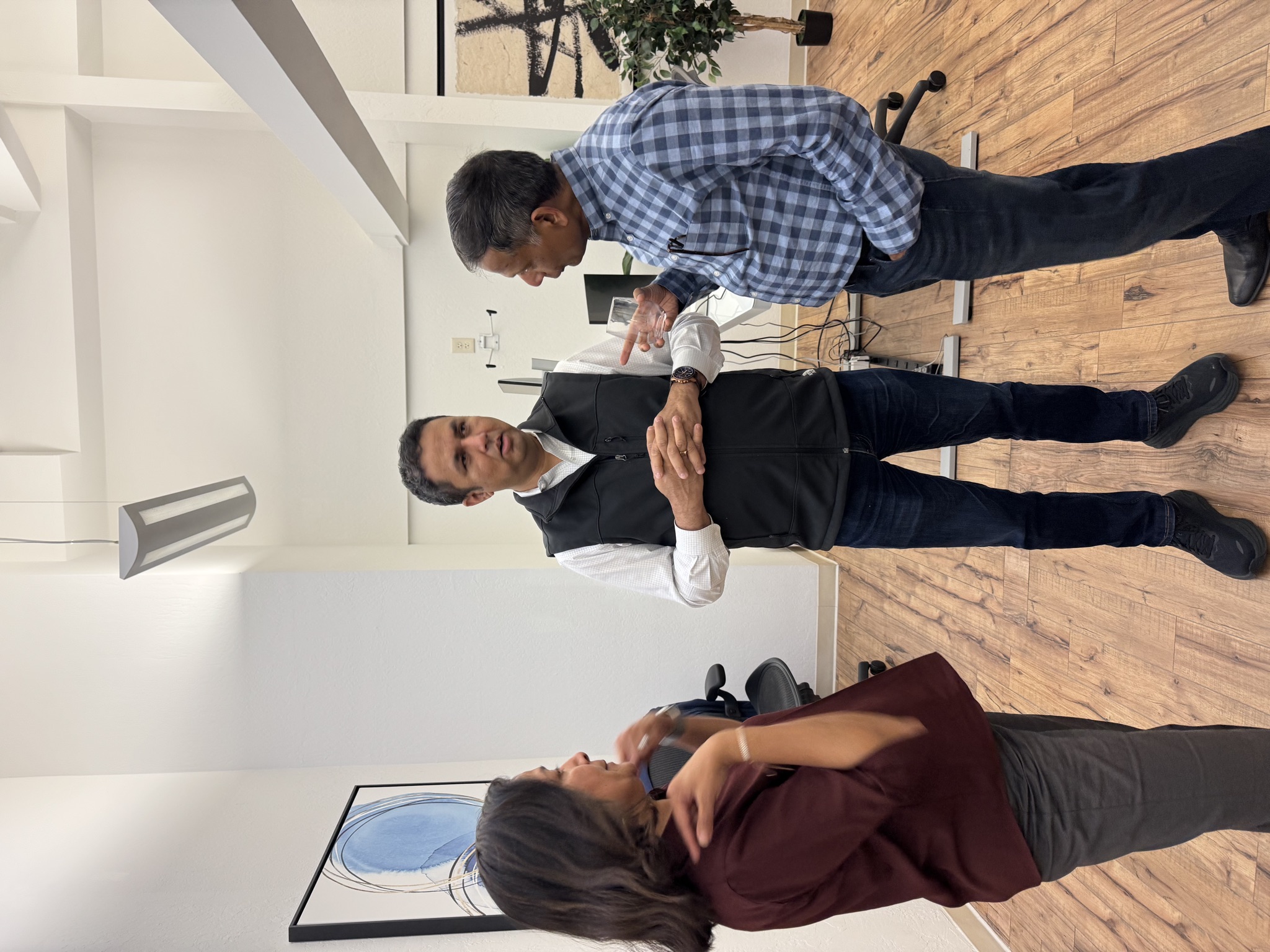 Three members in business casual attire conversing in a well-lit office with wooden floors and modern decor.