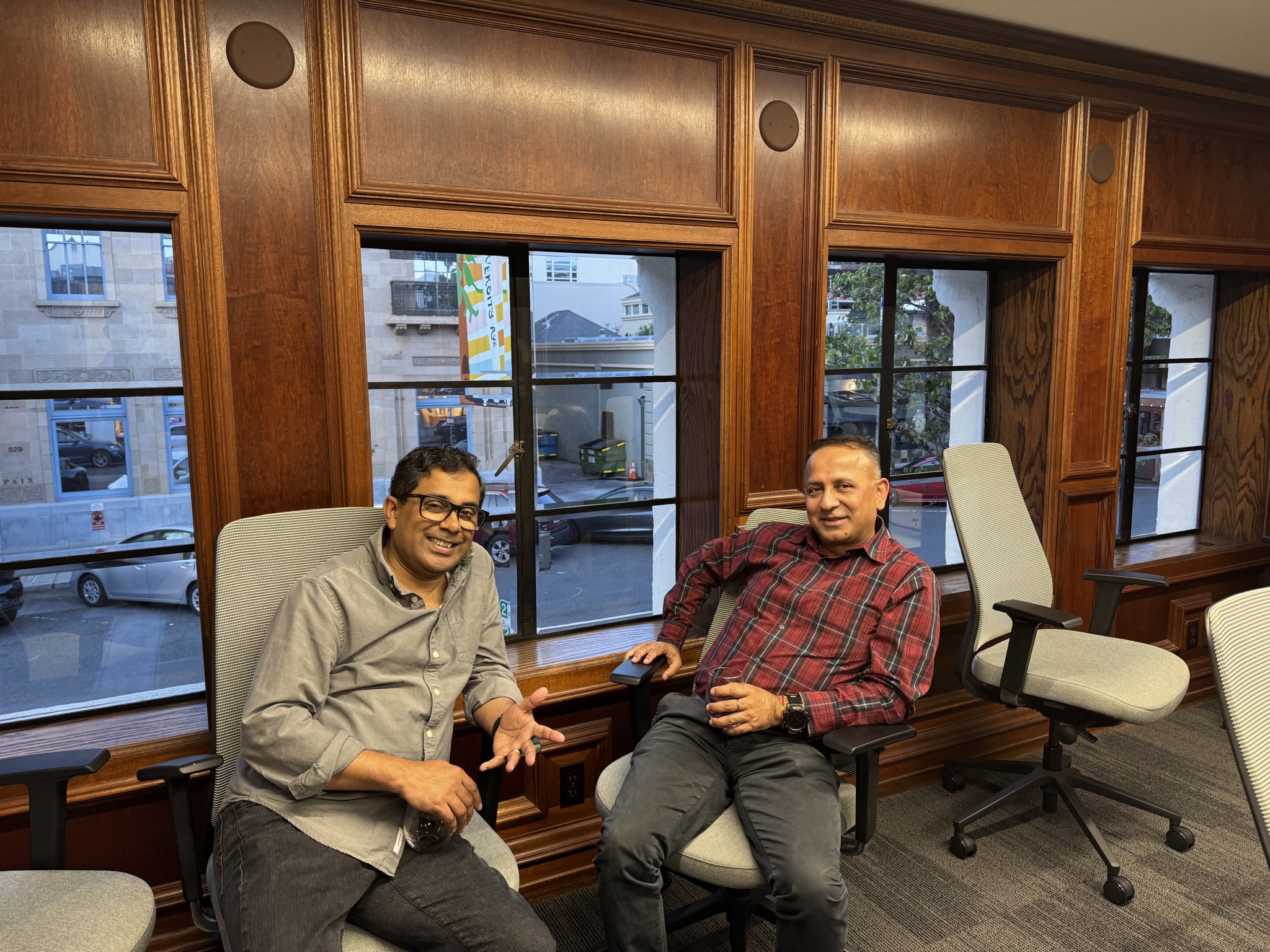 Two men sit in office chairs facing the camera in a room with wood-paneled walls and large windows overlooking a street.