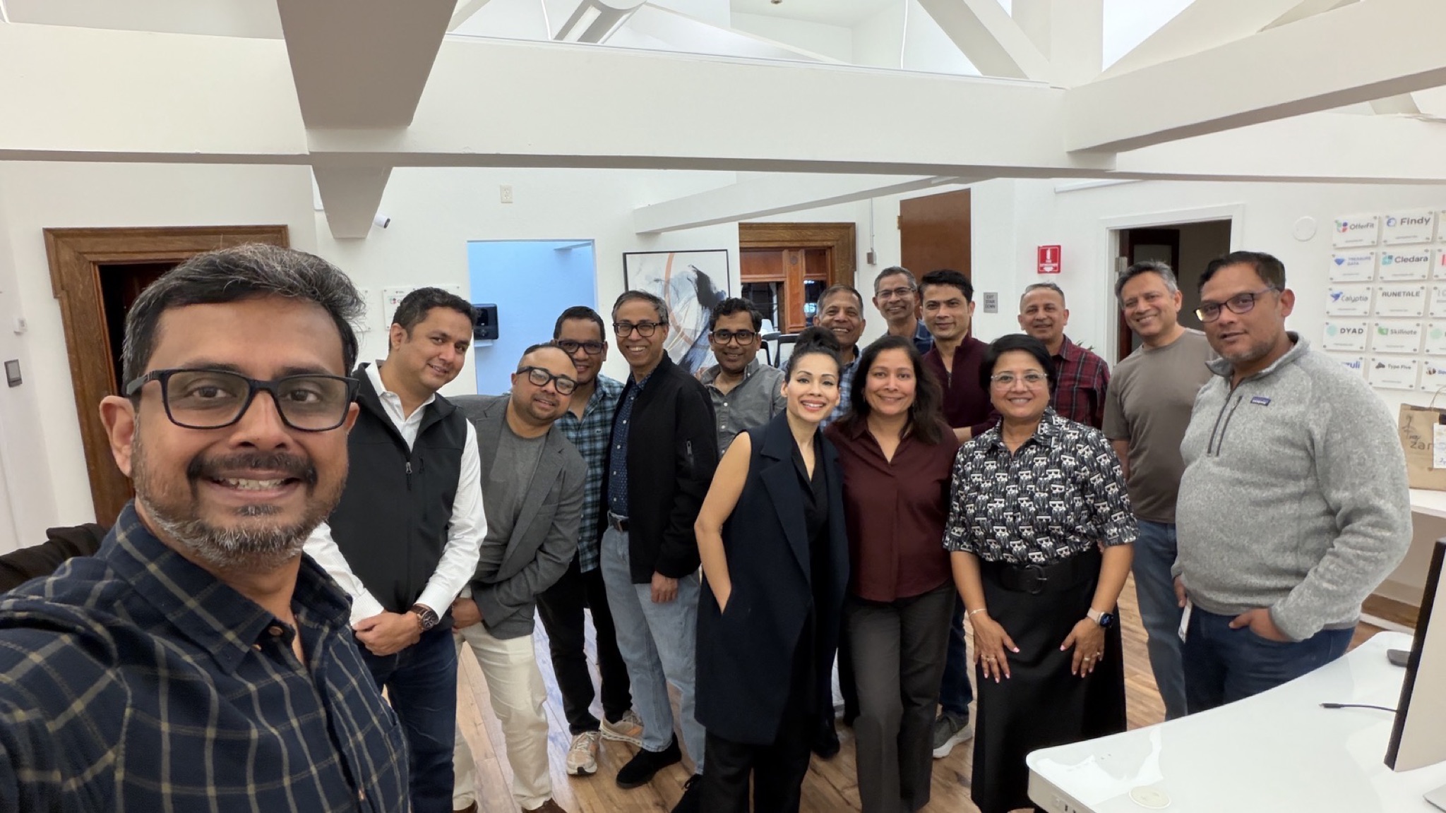 A group of approximately 15 diverse adults stand in an office setting, some smiling for the camera. They are dressed in casual and business attire.