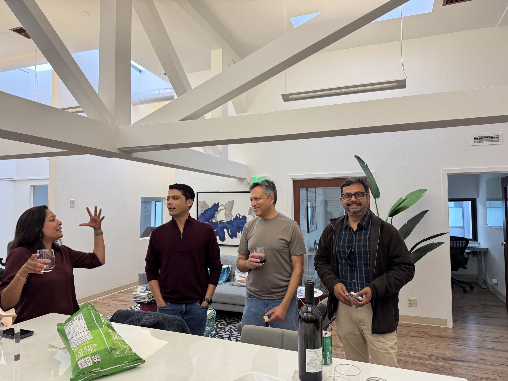 Four individuals stand in a well-lit, modern office space with exposed beams, holding drinks and engaging in conversation.