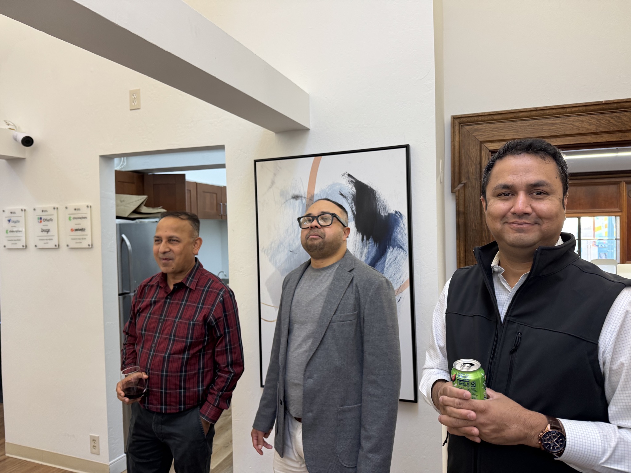 Three men in a well-lit room with artwork and signage. One man holds a glass of red wine, another a can of soda.