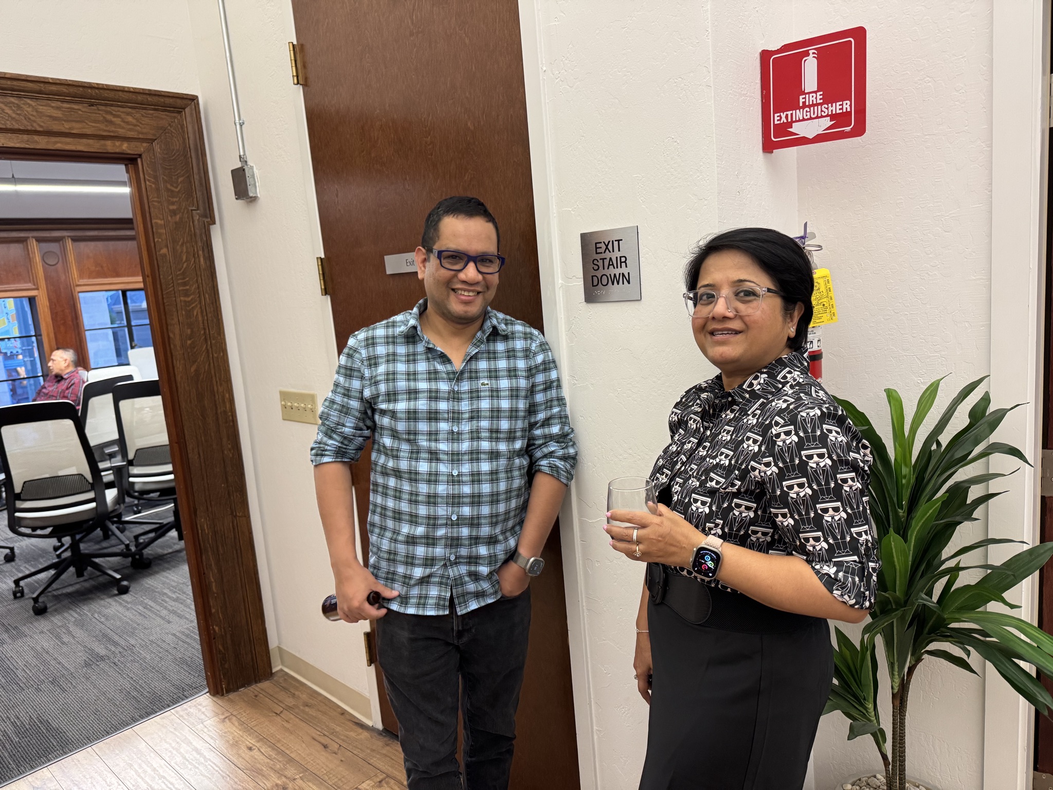 Two smiling members stand in an office hallway near a door, a fire extinguisher, and a plant. One holds a drink.