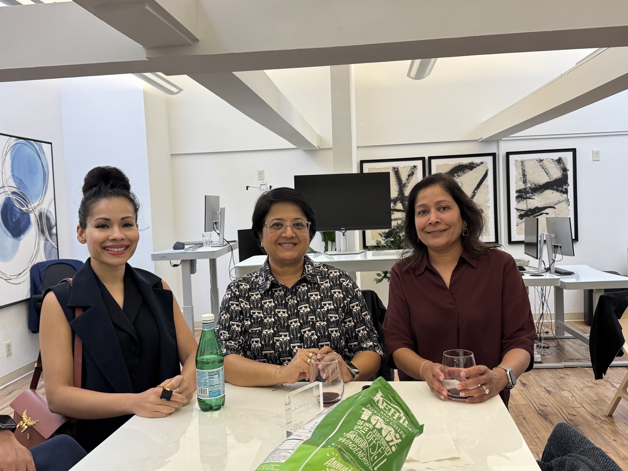 Three women sit at a white table in an office setting, smiling. One holds a glass of red wine, another a bottle of water, and a bag of chips is on the table.