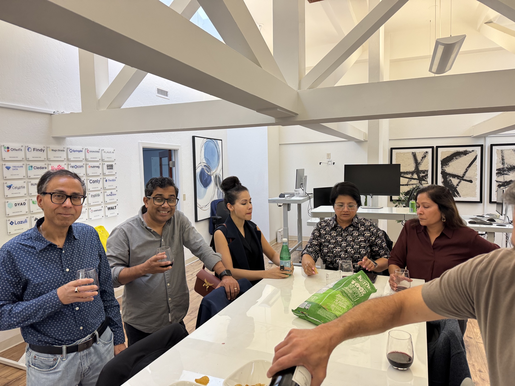 A group of six people of diverse backgrounds are gathered around a white table, holding drinks and snacks in a brightly lit modern office.
