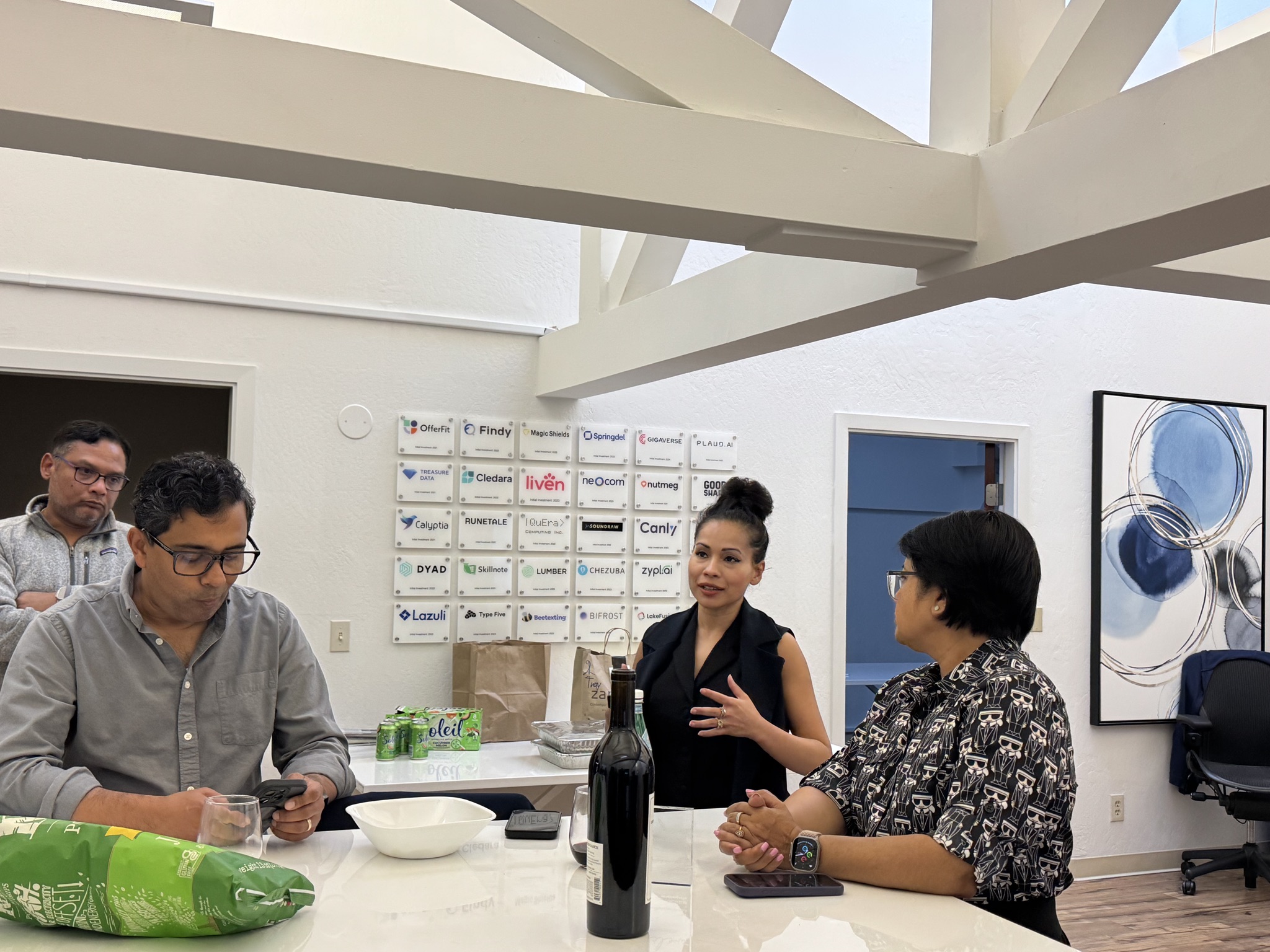 Four people gathered around a white table in a modern office setting, with a wall of company logos behind them.