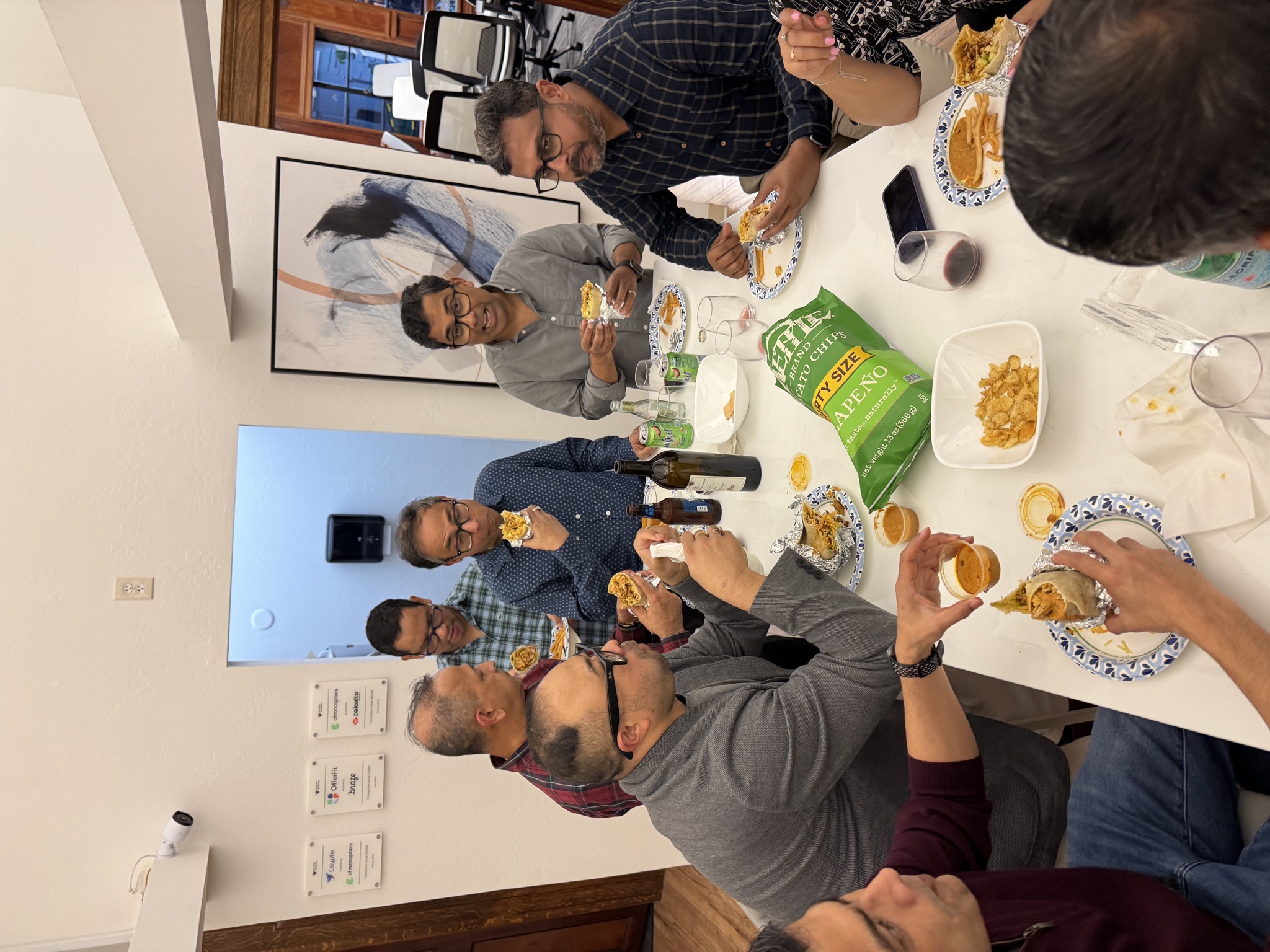 A group of men sit around a white table, eating burritos and chips, with drinks and bottles visible.